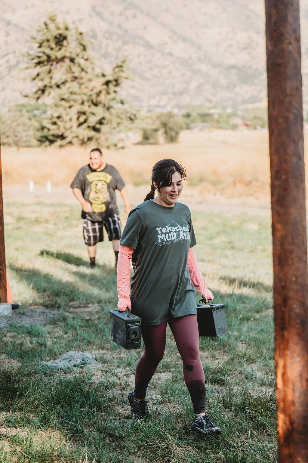Two people outdoors, one carries metal boxes labeled "Tenachapi Mud Run," with mountains in the background.