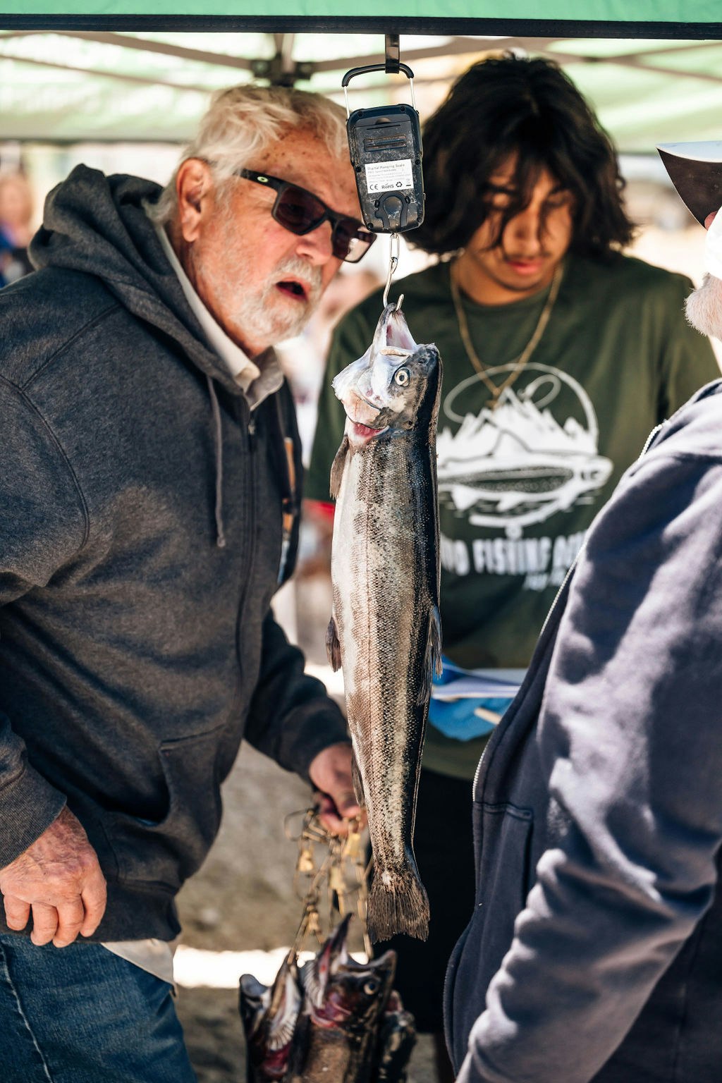 A man weighs a fish on a digital scale at a market while others watch.