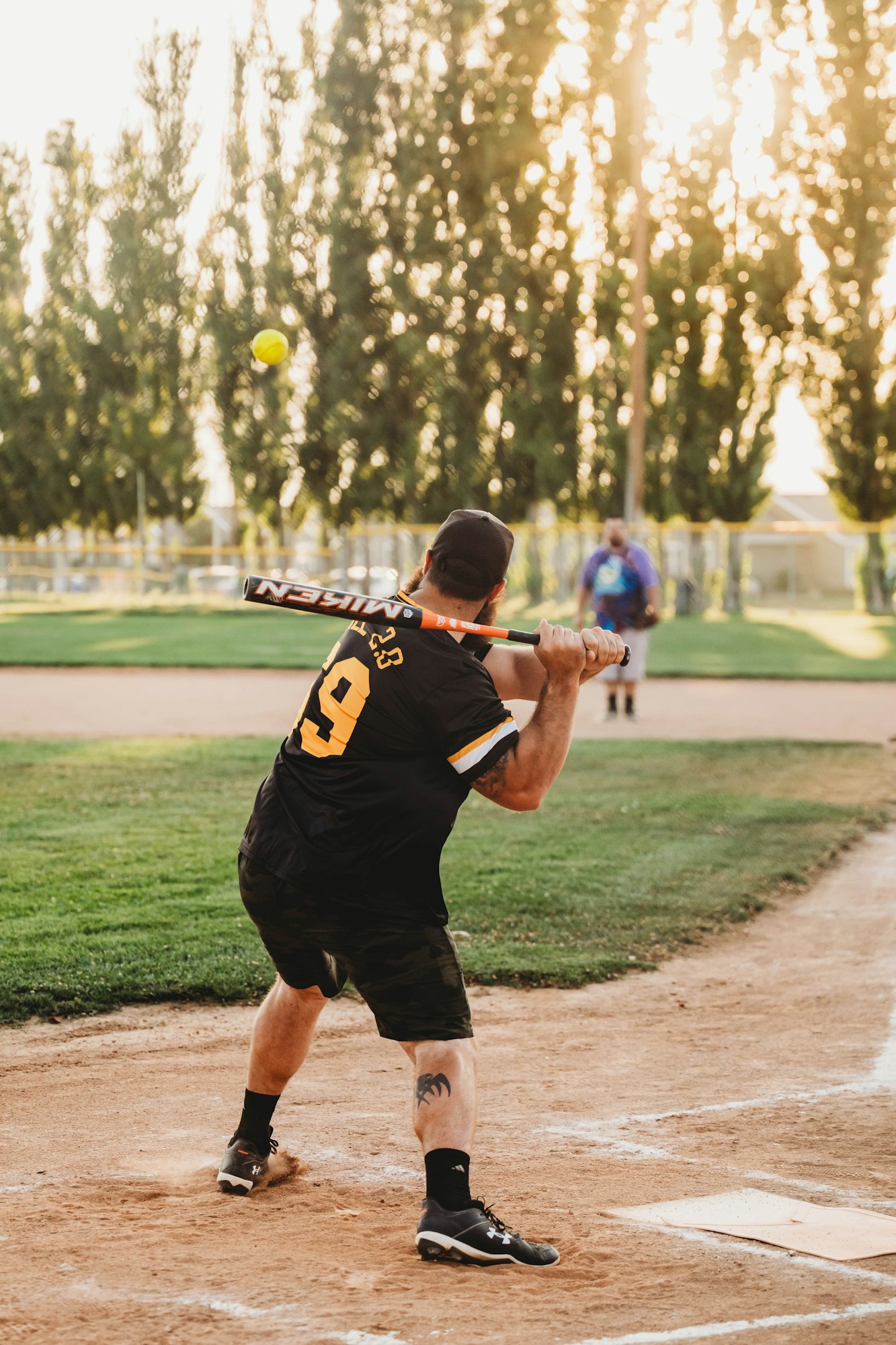 A player swinging a bat at a softball game, trees and sunlight in the background.