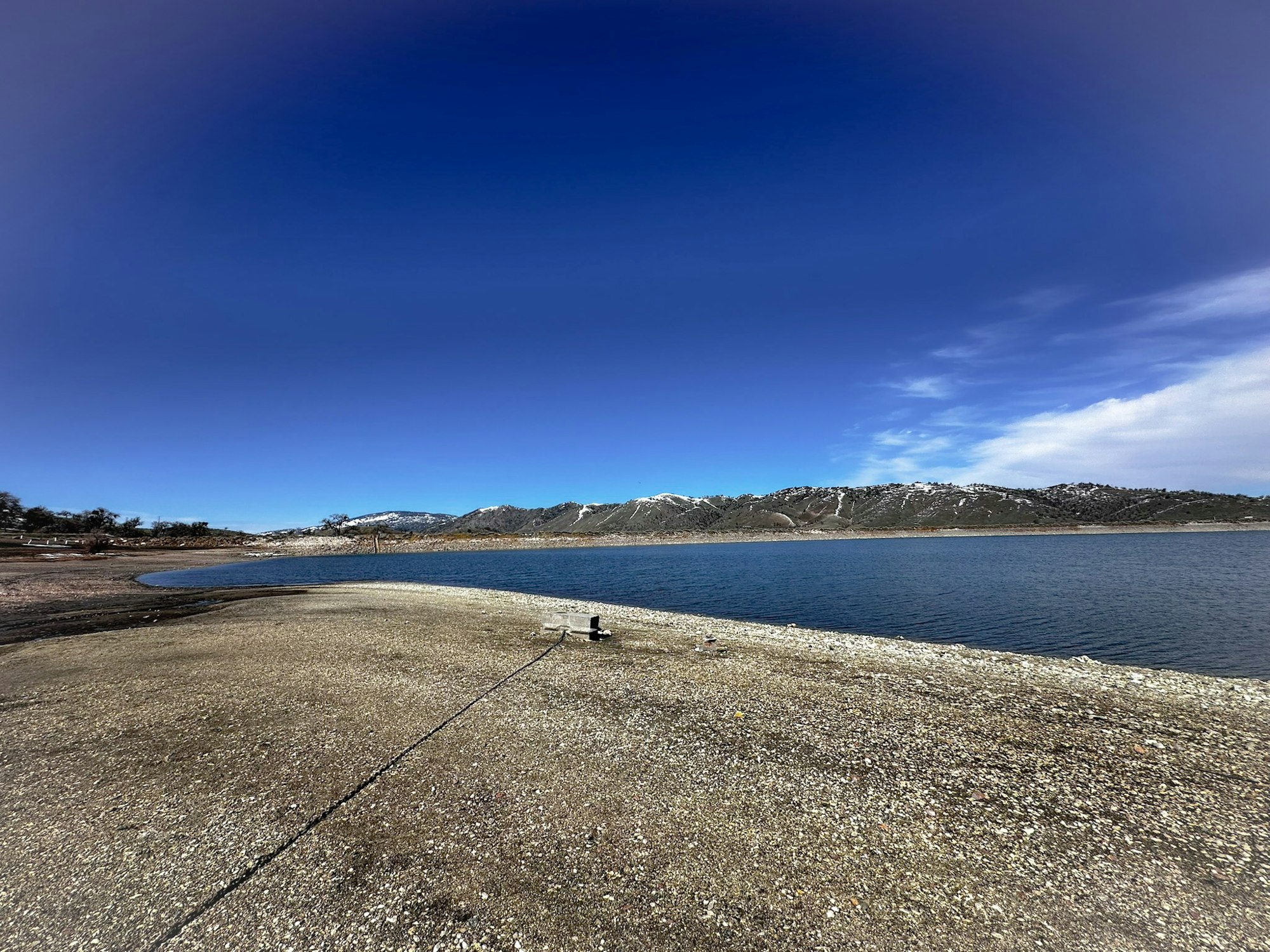 A clear blue sky, a calm lake, a curving shoreline, mountains in the distance, and a solitary bird on the ground.