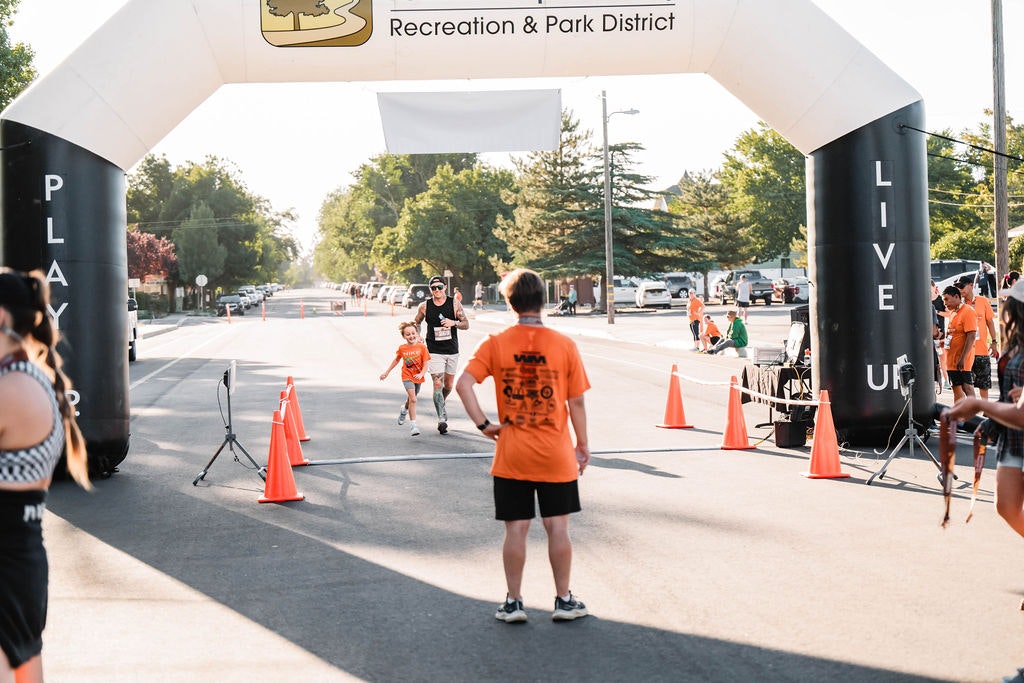 Runners crossing the finish line at a park district event, with people cheering and an inflatable arch overhead.