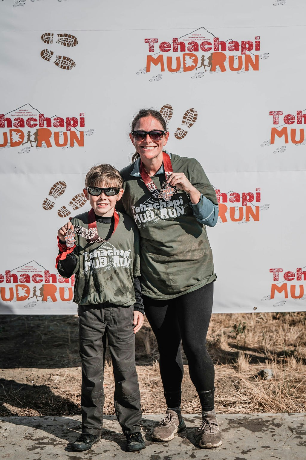Two people in muddy shirts with medals, posing in front of a "Tehachapi Mud Run" backdrop.