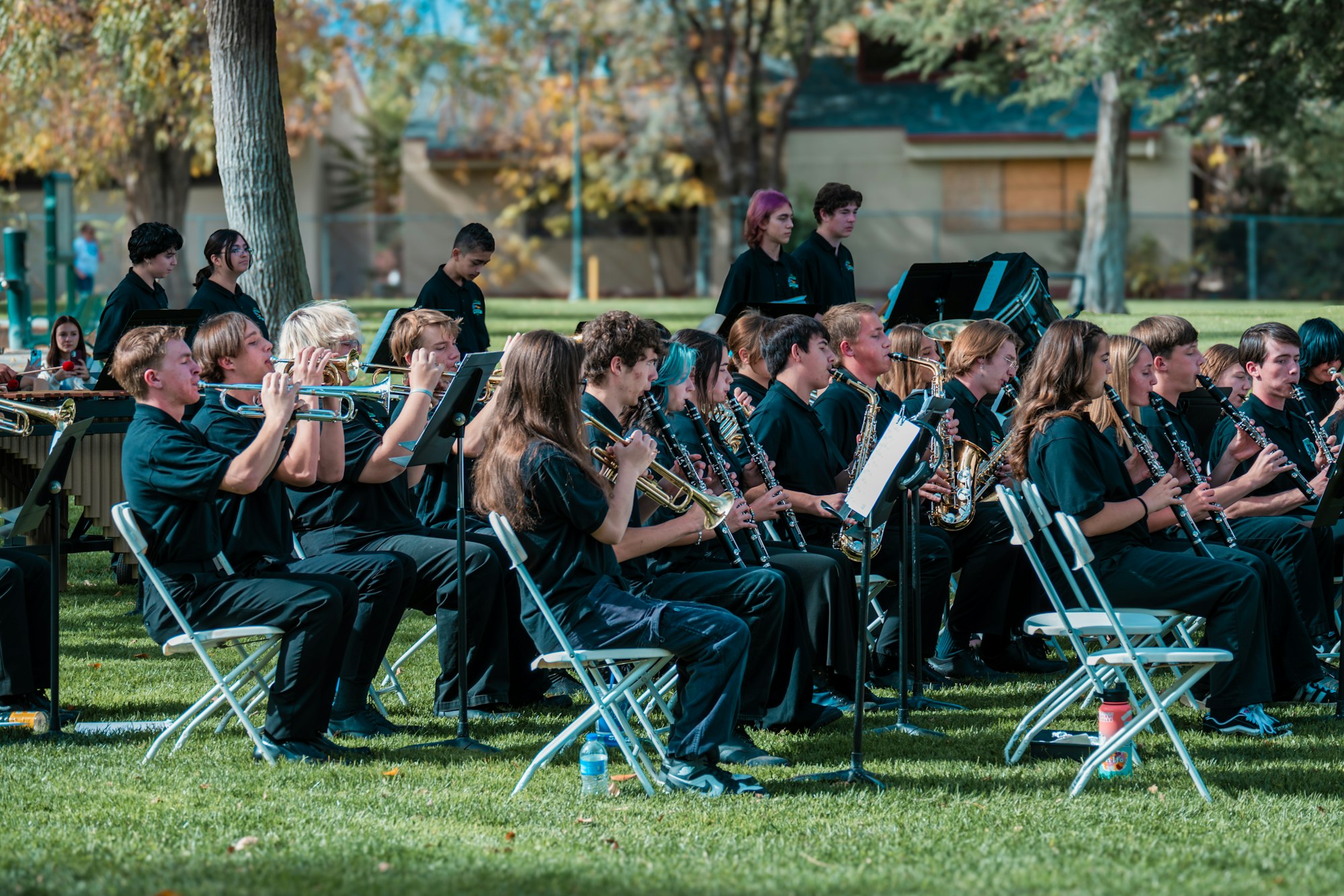 A seated band of musicians in uniform performs outdoors with various instruments like trumpets and clarinets.