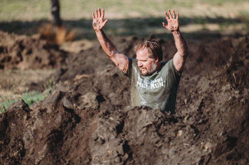 Person in a muddy trench with arms raised, participating in an outdoor event.