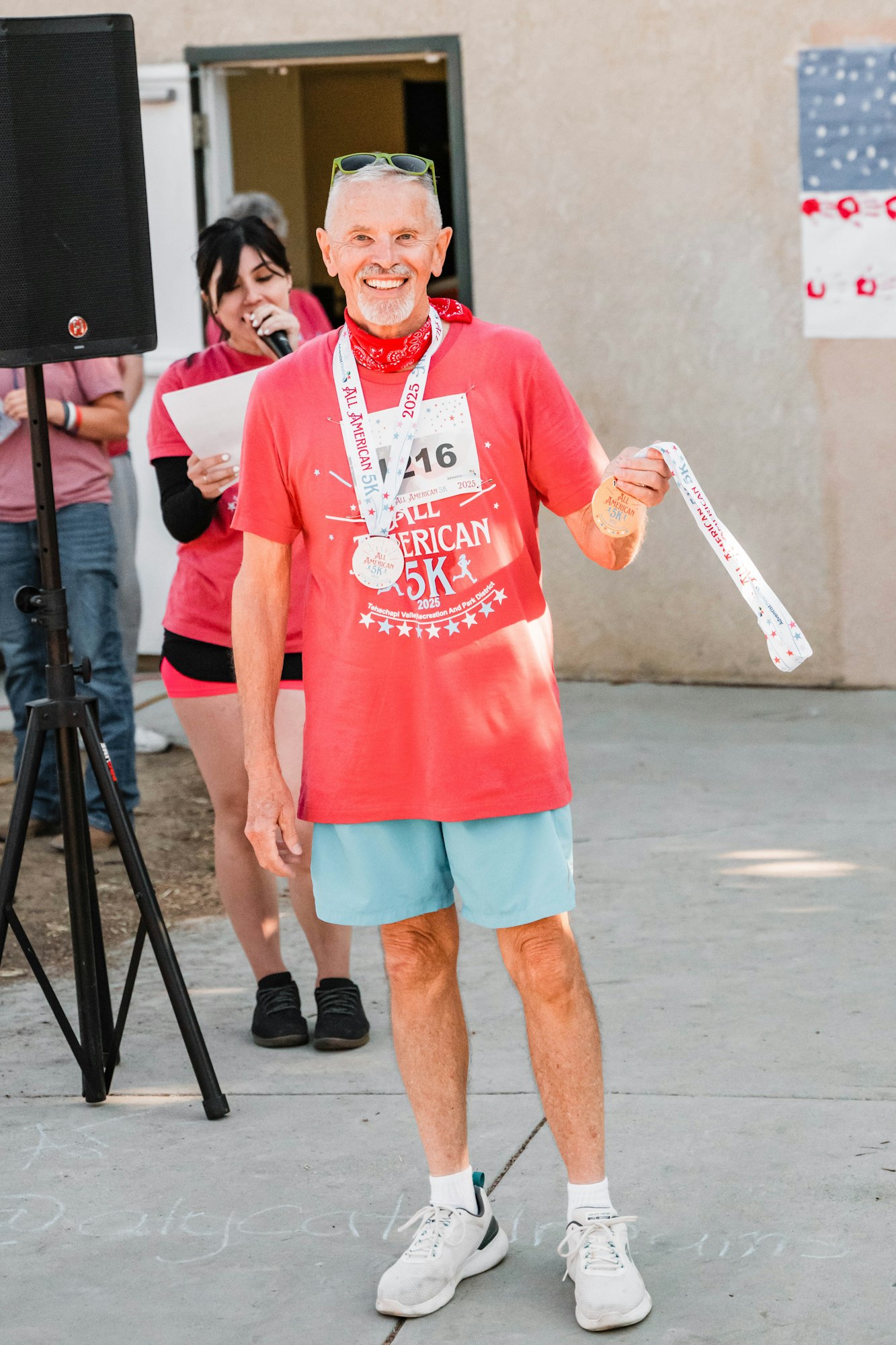 A man wearing a "5K" shirt holds a medal, smiling. A woman is speaking into a microphone behind him.