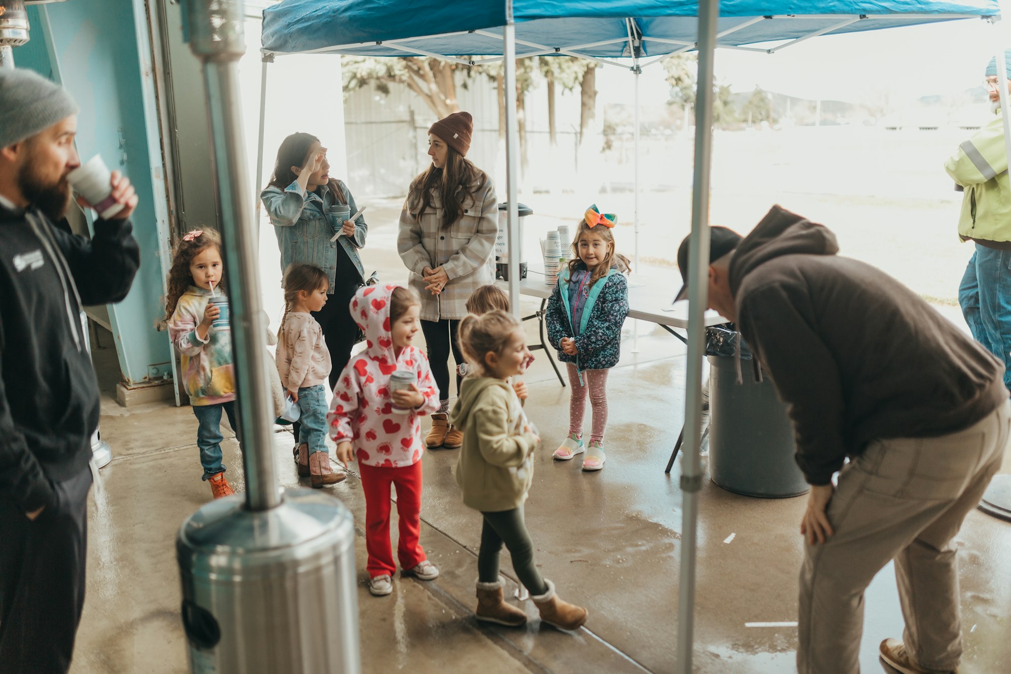 Group of people, including children, gathered under a canopy, possibly at a casual outdoor event.