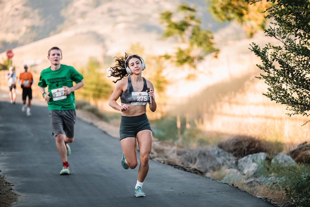 People running on a paved path in warm weather, with a woman leading and wearing headphones.
