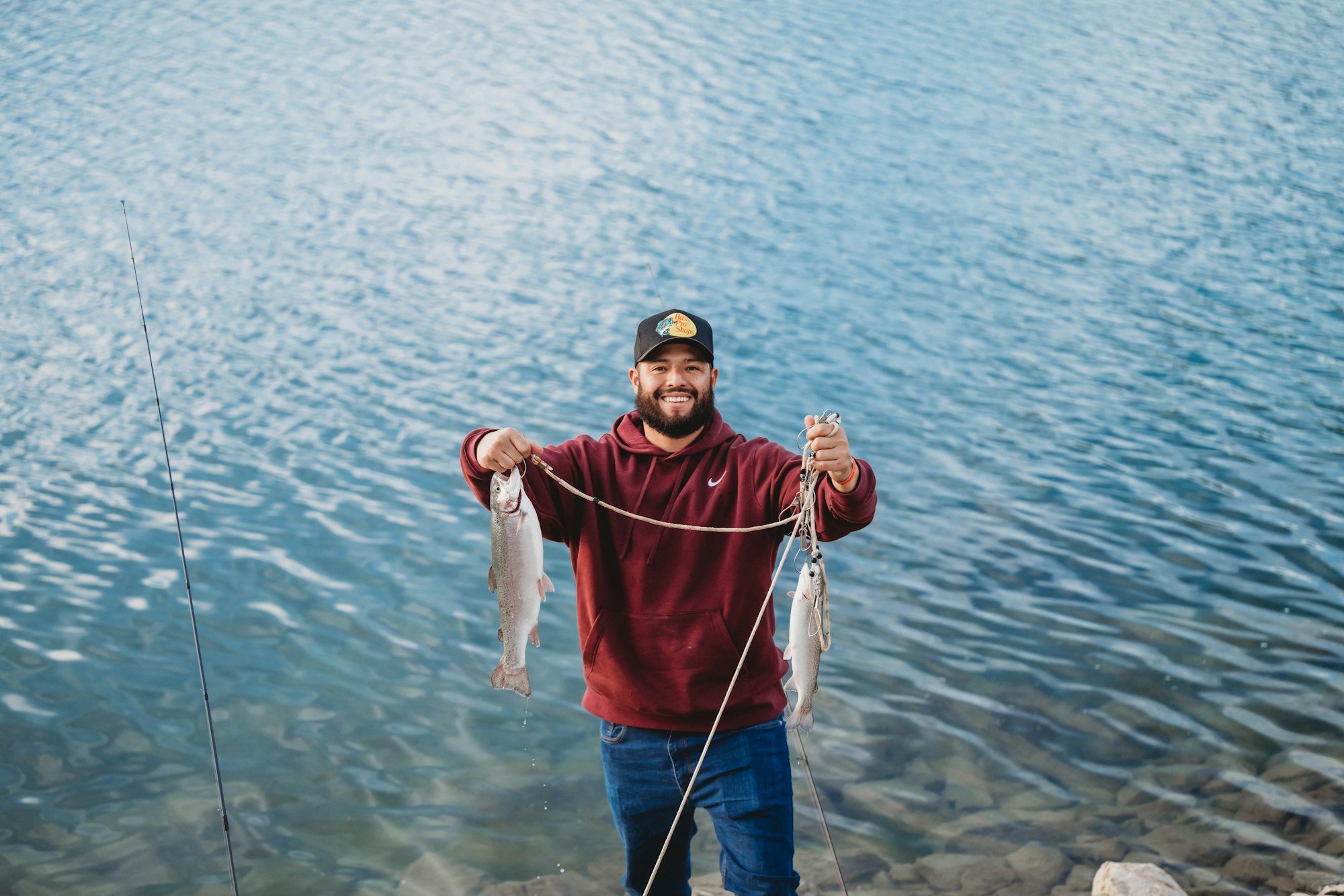 A person smiles holding two freshly caught fish by a lake, wearing a maroon hoodie and cap.