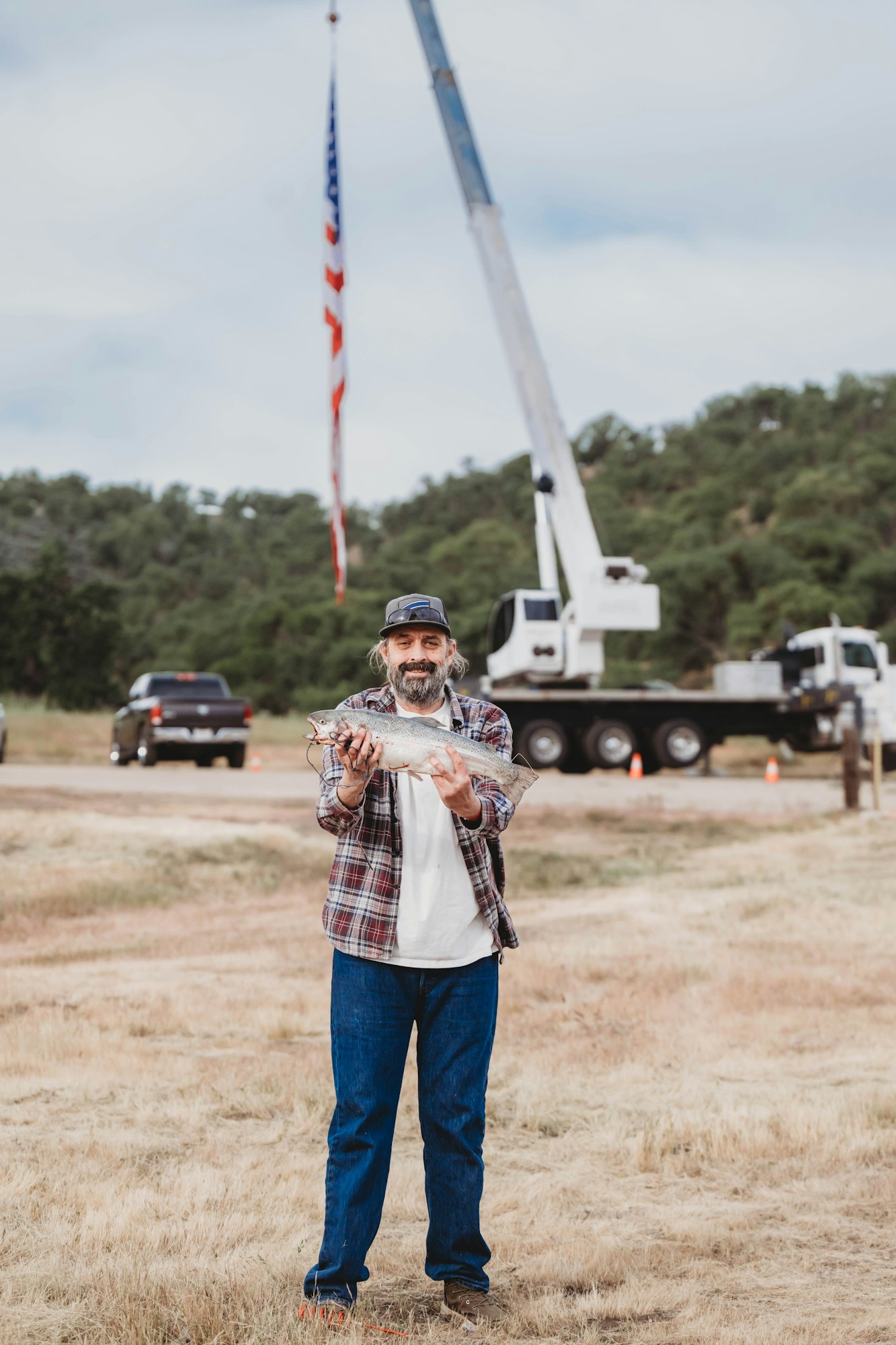 A person holding a fish stands in front of a crane with a U.S. flag, in a grassy field.