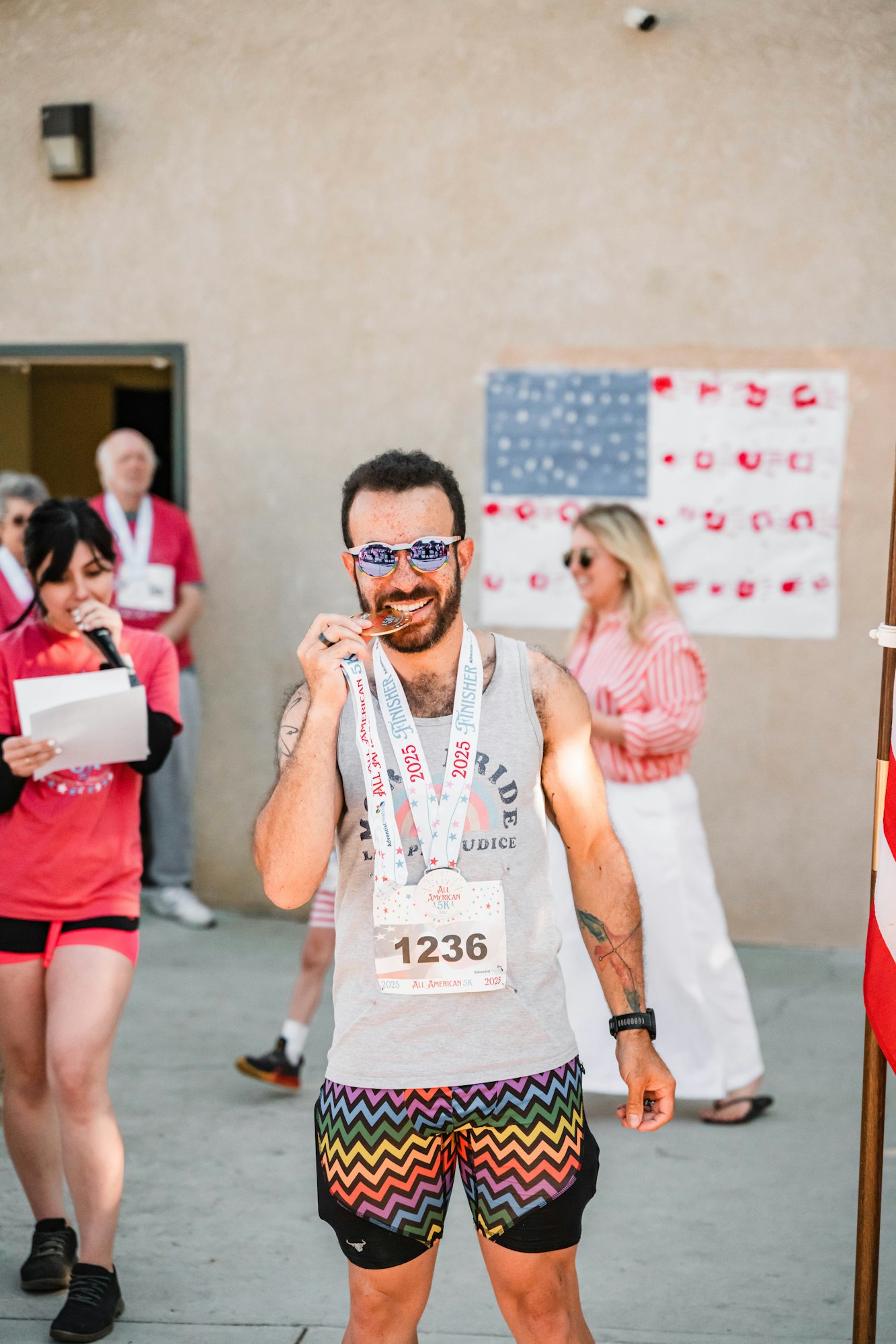 A person in colorful shorts and sunglasses celebrates a race finish with medals, surrounded by people and an American-themed backdrop.