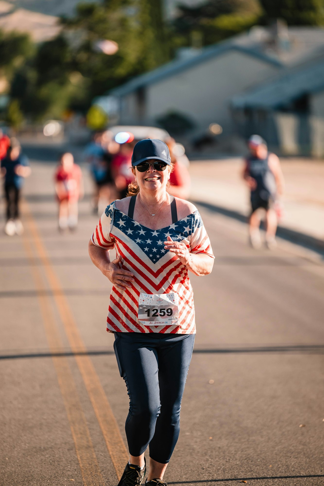 A woman running in a race, wearing a patriotic shirt and number 1259 bib, with others in the background.