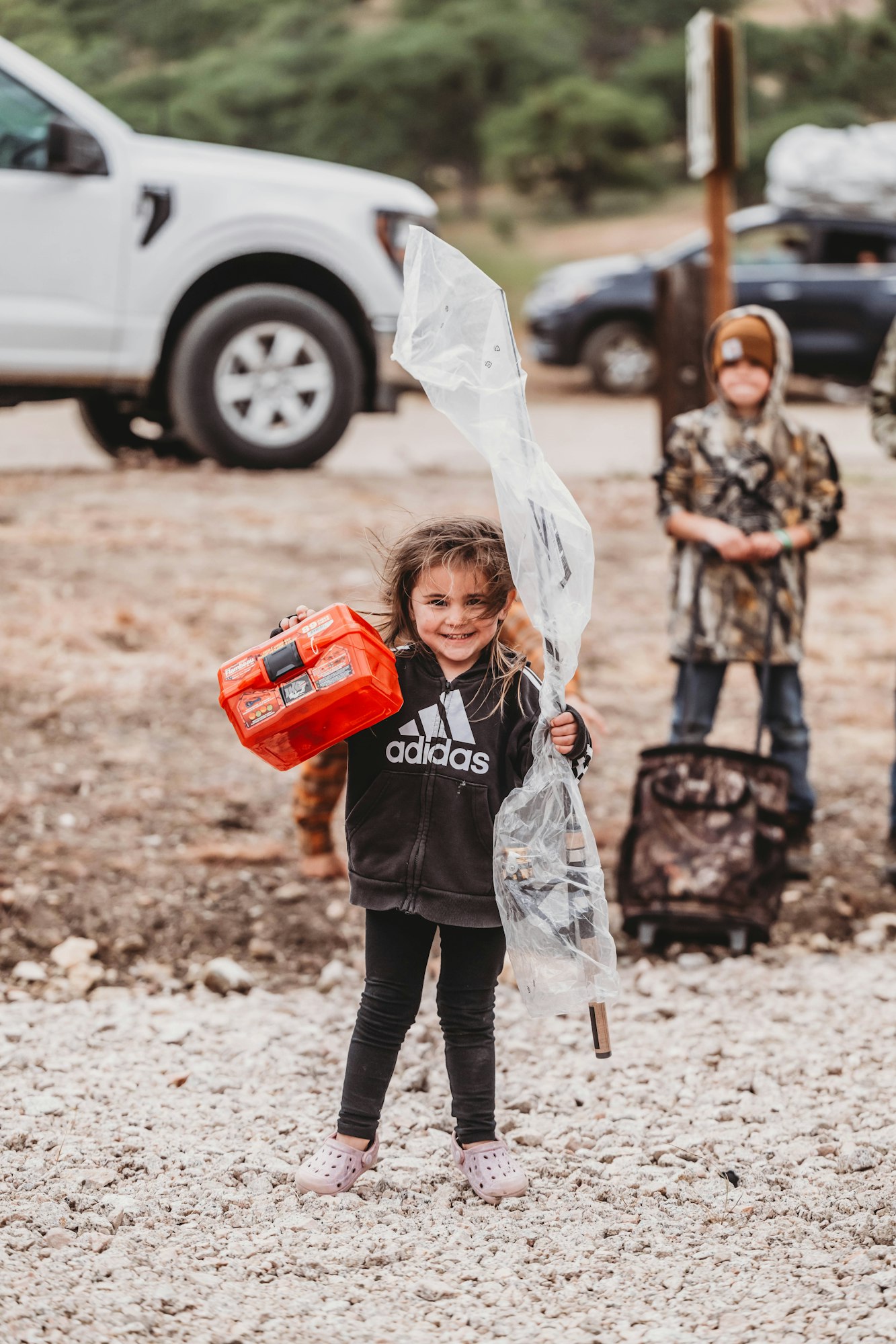 A smiling child holding a red toolbox and plastic-wrapped item, outdoors with vehicles and another child in the background.