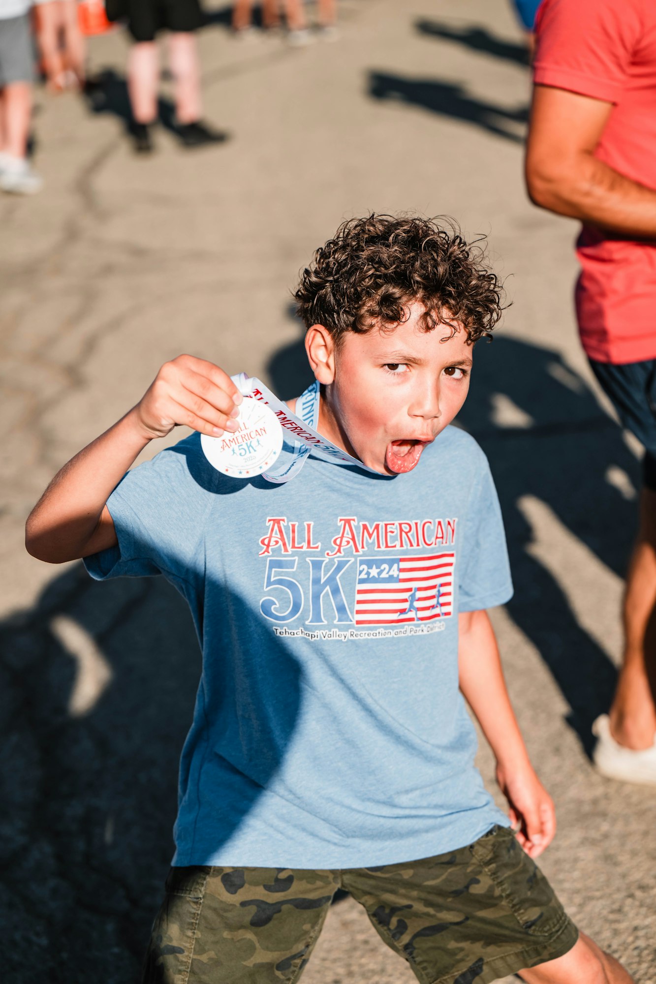 Child celebrating with a medal, wearing a blue "All American 5K" shirt and camouflage shorts, surrounded by people outdoors.