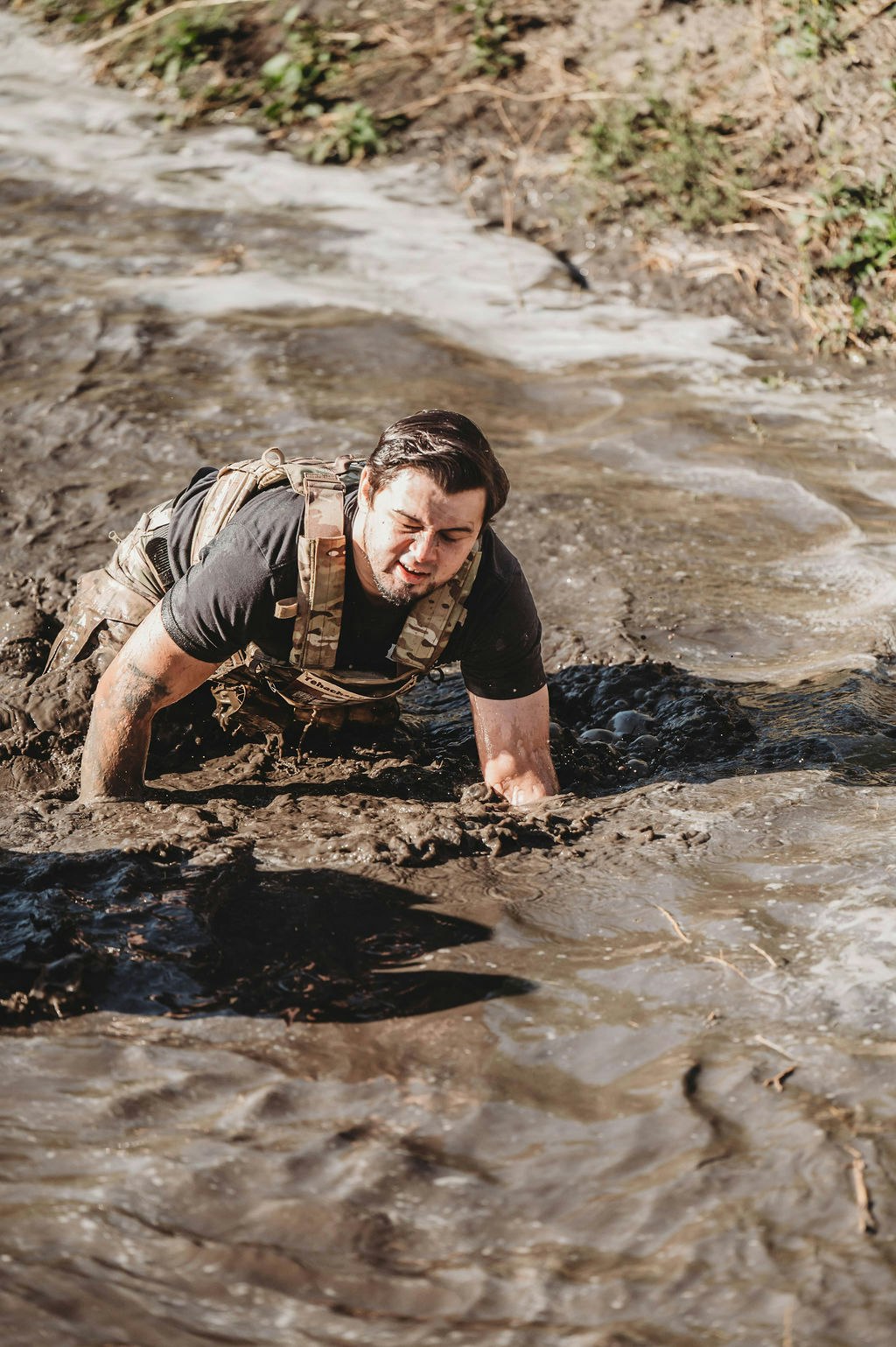 A person in tactical gear is crawling through muddy water outdoors.