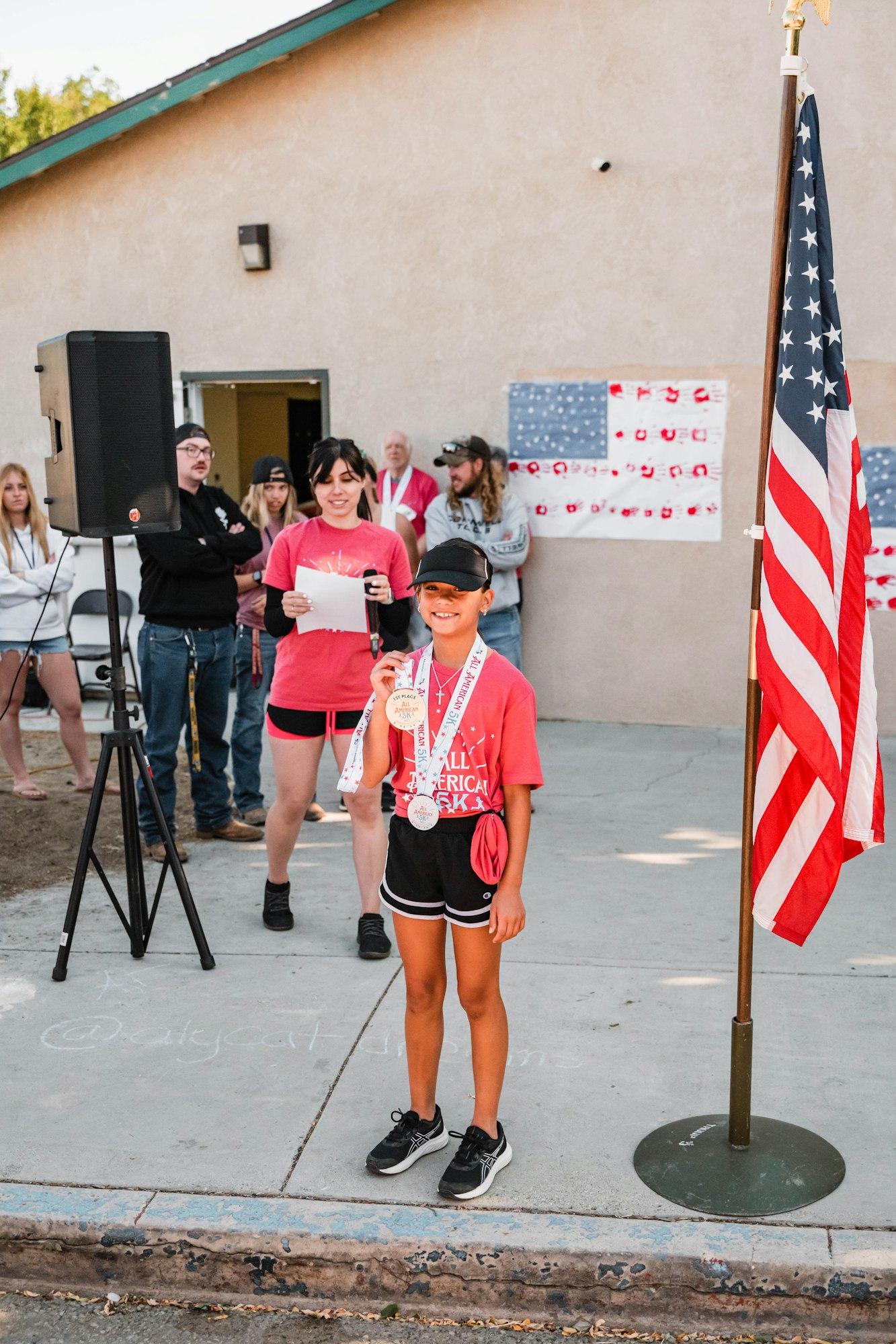 A young person with medals smiles near a U.S. flag, with a group of onlookers and a speaker in the background.