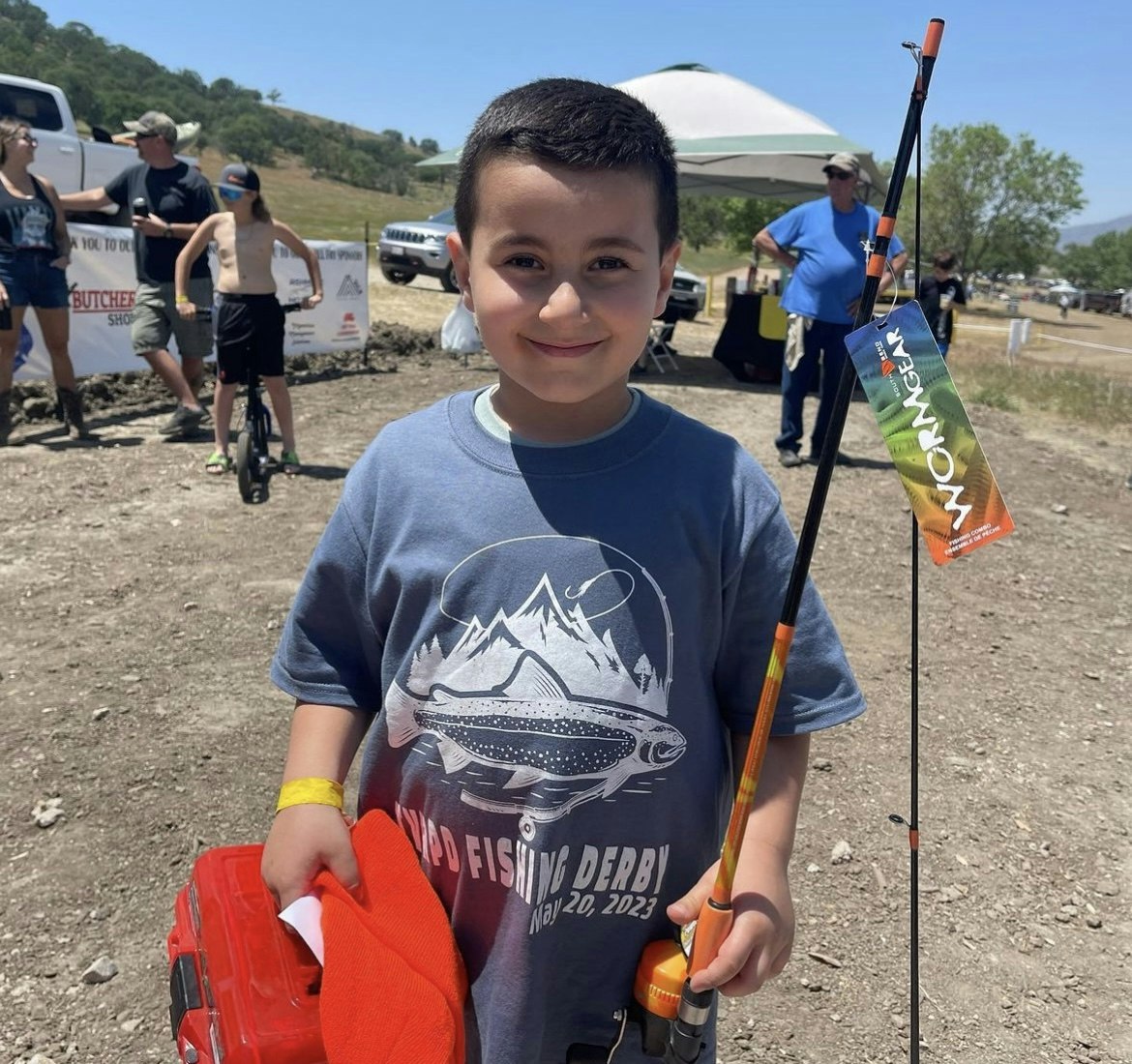 A smiling boy holding a fishing rod and a tackle box at an outdoor fishing event.