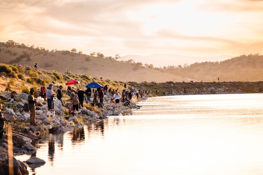 People by a lakeside during a golden sunset, some with umbrellas, in a serene landscape setting.
