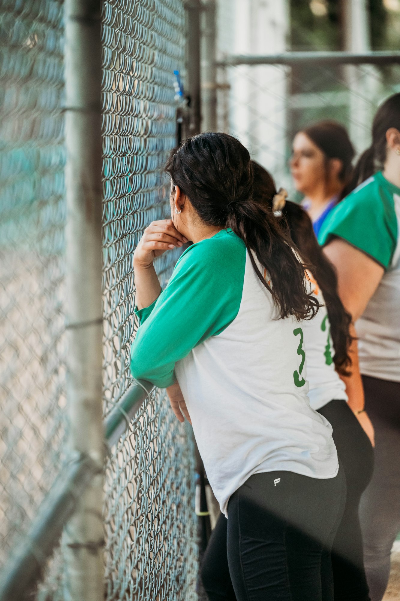 A person in a sports jersey leaning against a chain-link fence, possibly watching a game or awaiting their turn to play.