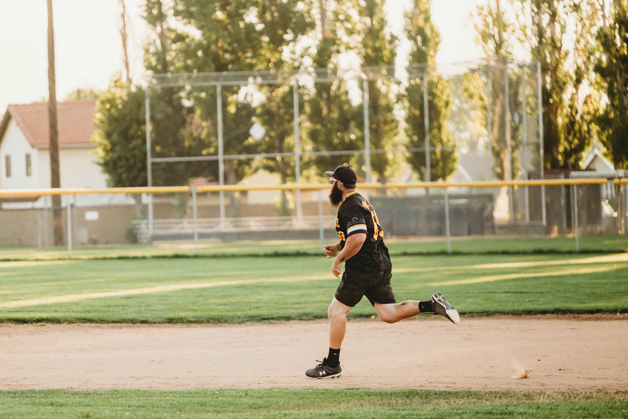 A person running on a baseball field with back to the camera, in sports attire and cap, with chain-link fences and trees in background.