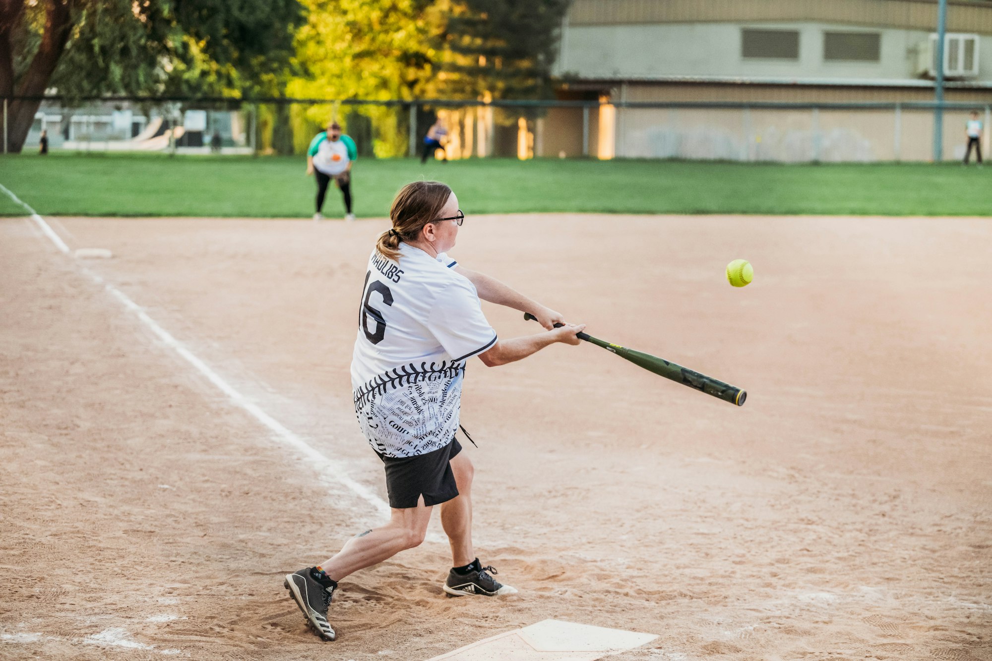 A person playing softball is swinging at a pitch, with an outfielder visible in the background.
