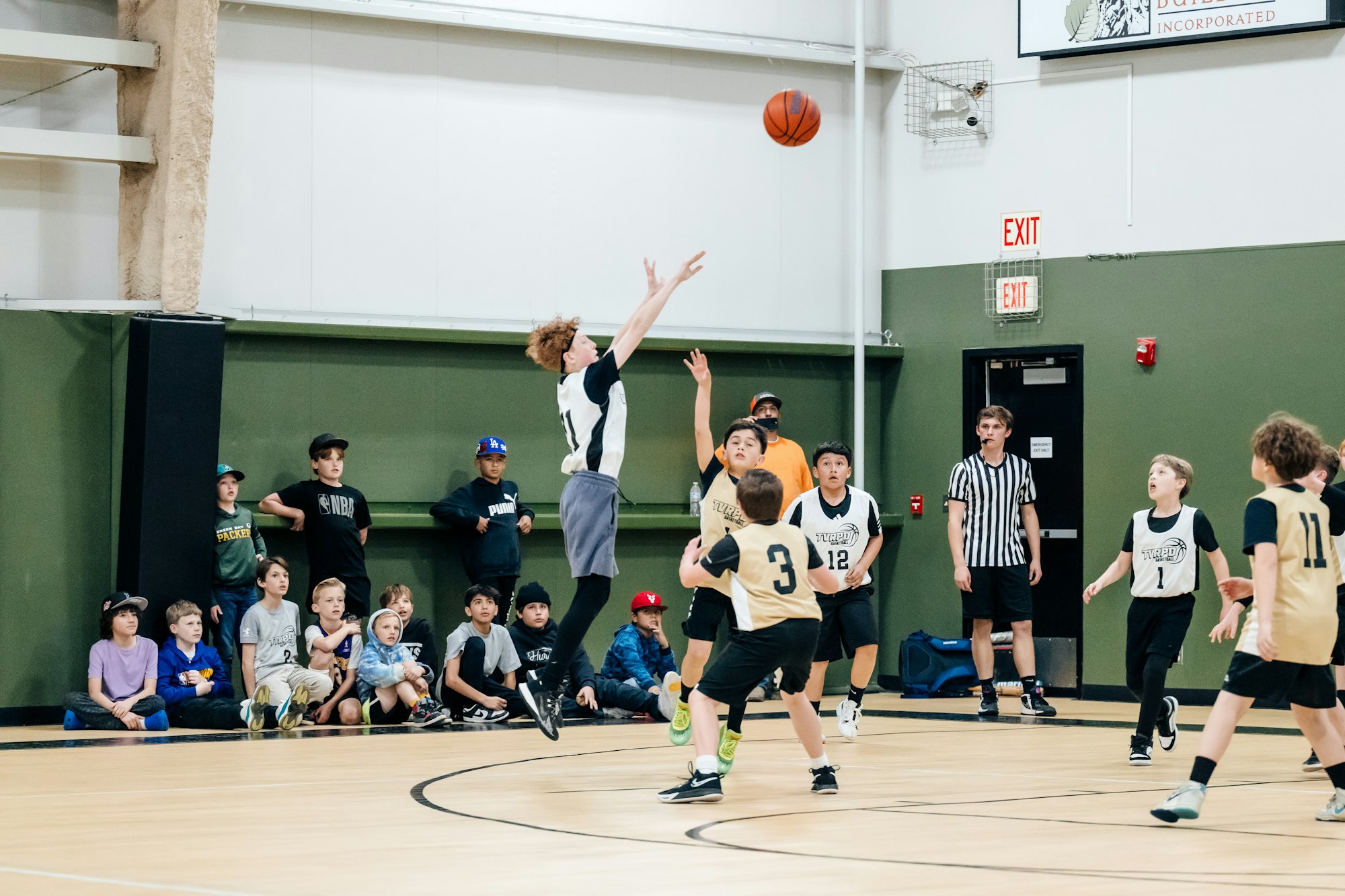 Youth basketball game with a player shooting, kids watching, and a referee observing.