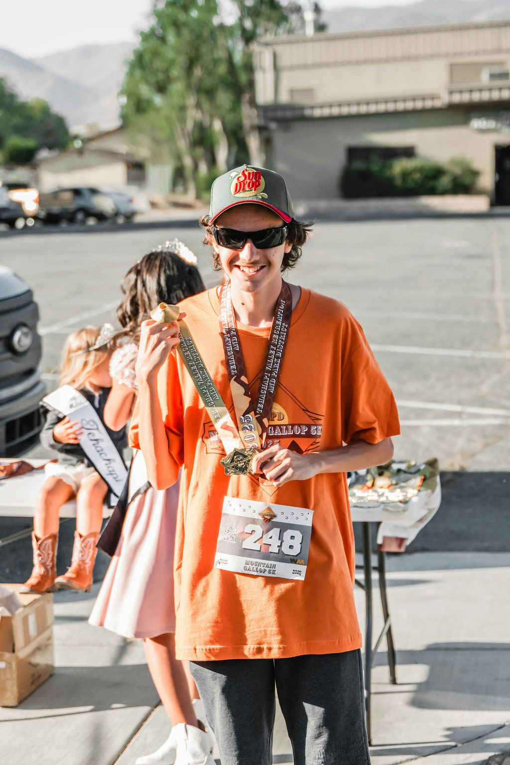 A person in an orange shirt, wearing sunglasses and a hat, holding a medal with a race bib number 248.