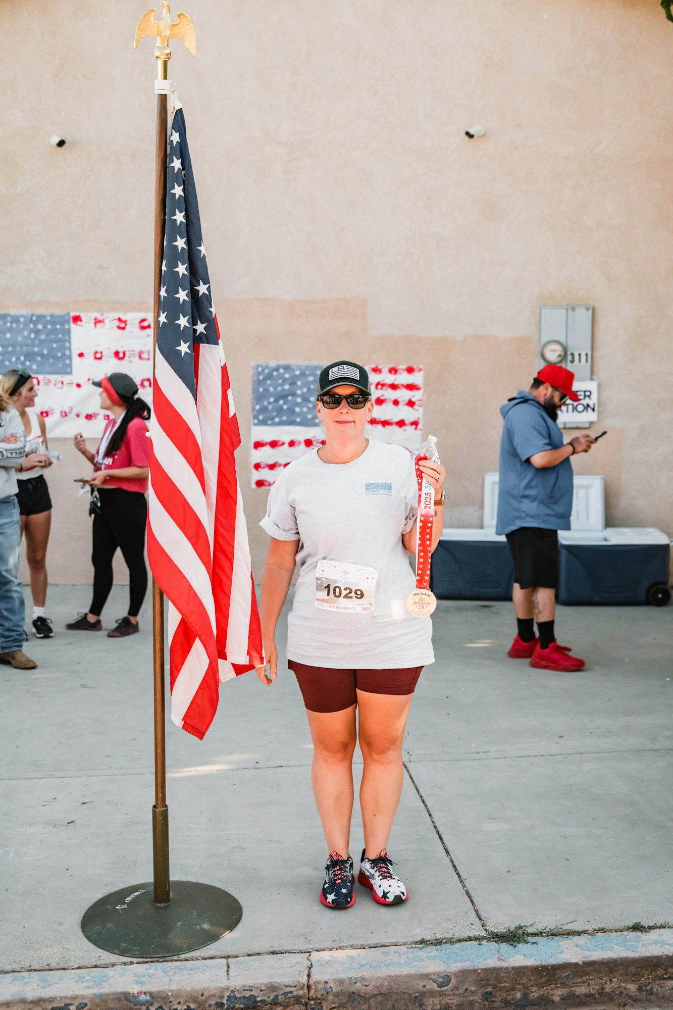 A person in running gear holding a medal, standing near a flagpole with the American flag, in front of a wall with flag art.