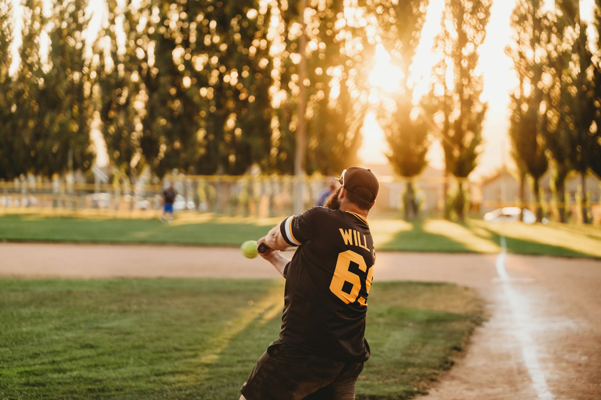 A person swings a bat at a softball on a field at sunset.