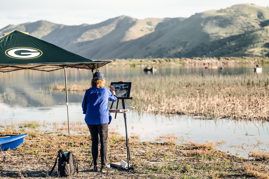 Person painting by a lake under a sports team canopy, with hills in the background and people on boats in the water.
