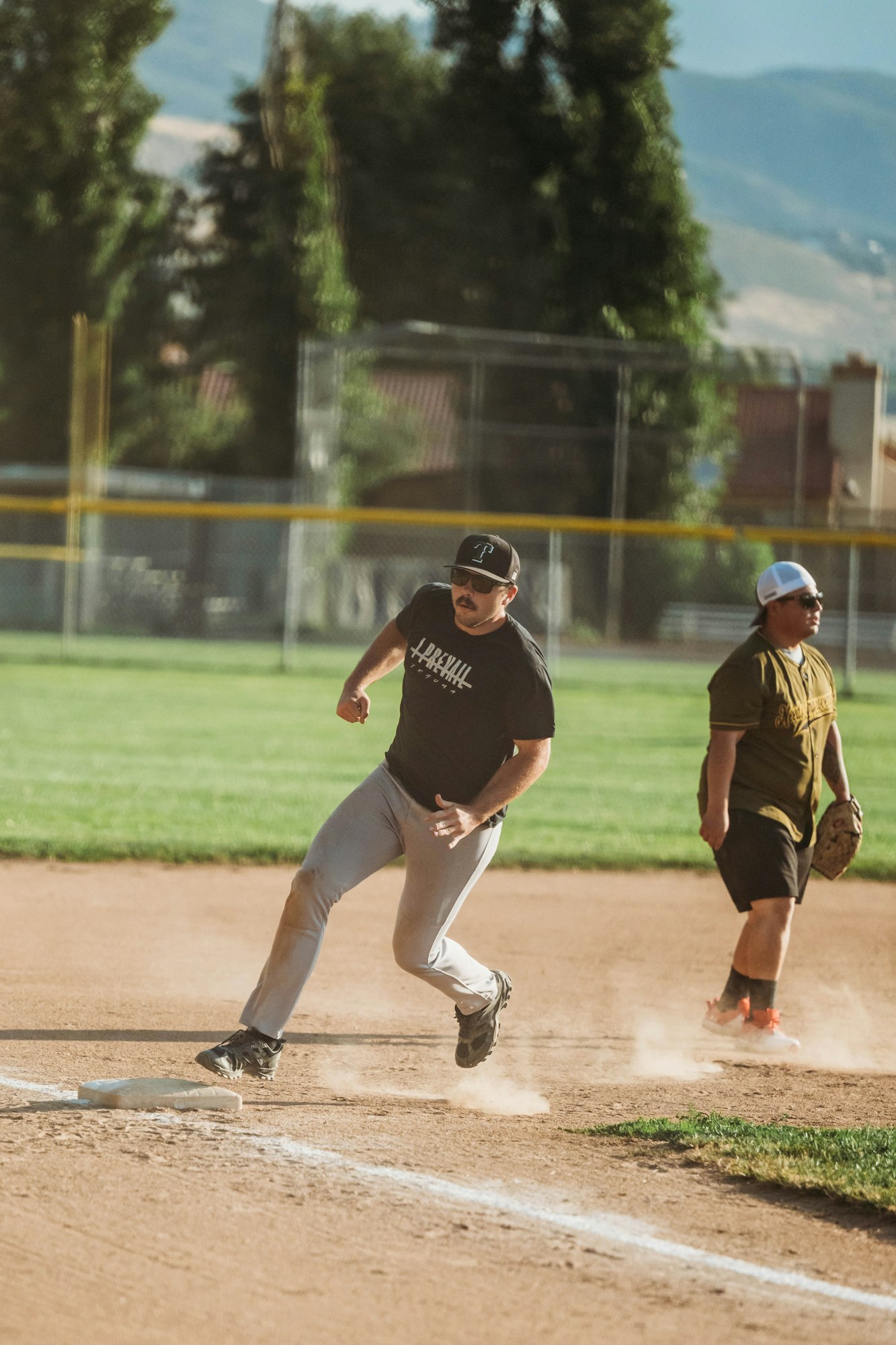 Two men playing baseball, one running bases, the other fielding, with a scenic backdrop.