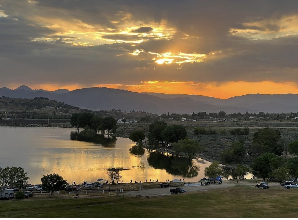 A scenic sunset with golden hues over a lake, mountains in the backdrop, and silhouetted trees reflecting on water. People are visible near parked cars.