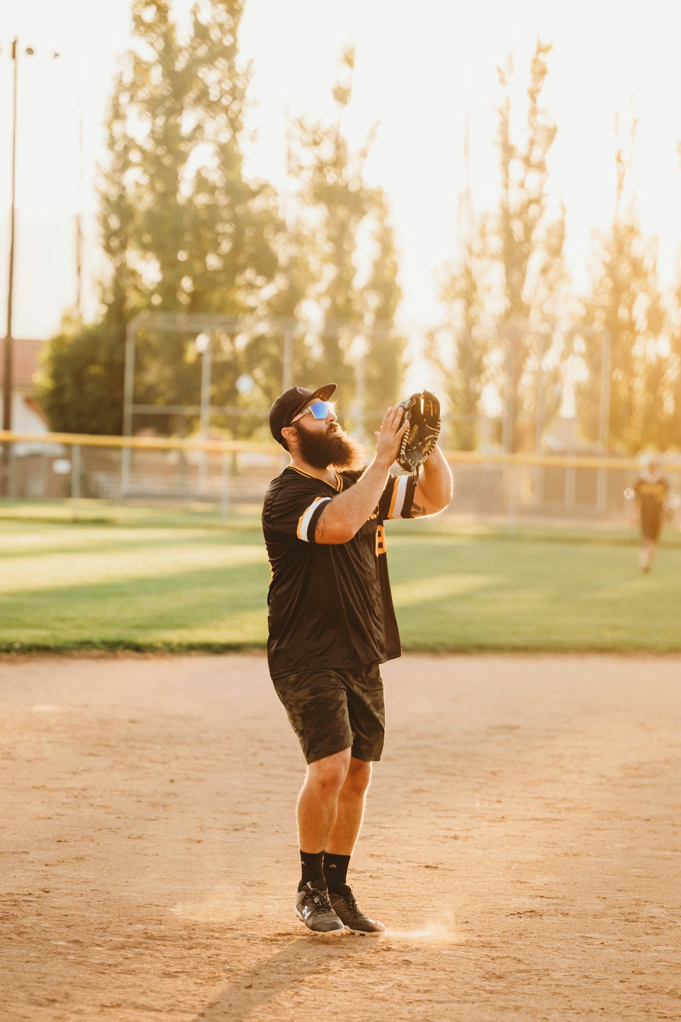 Man with a beard, wearing sunglasses, cap, and sports attire, catching a ball with a baseball glove.