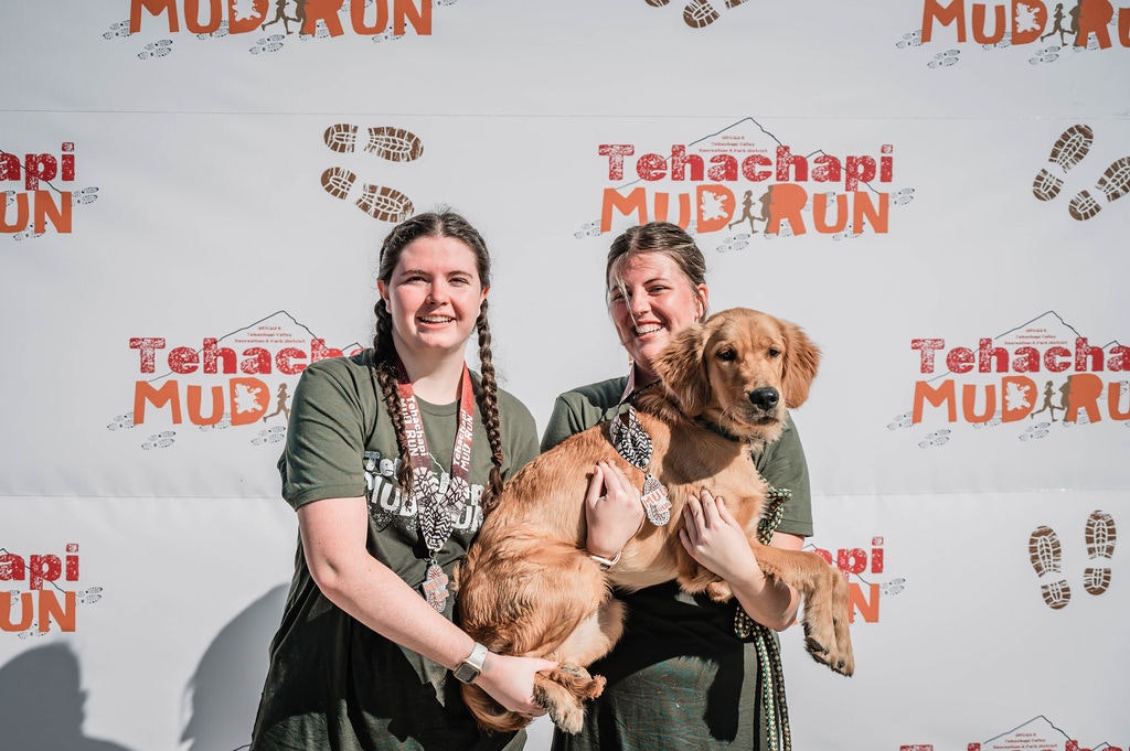 Two people holding a dog in front of a "Tehachapi Mud Run" backdrop, smiling and wearing medals.