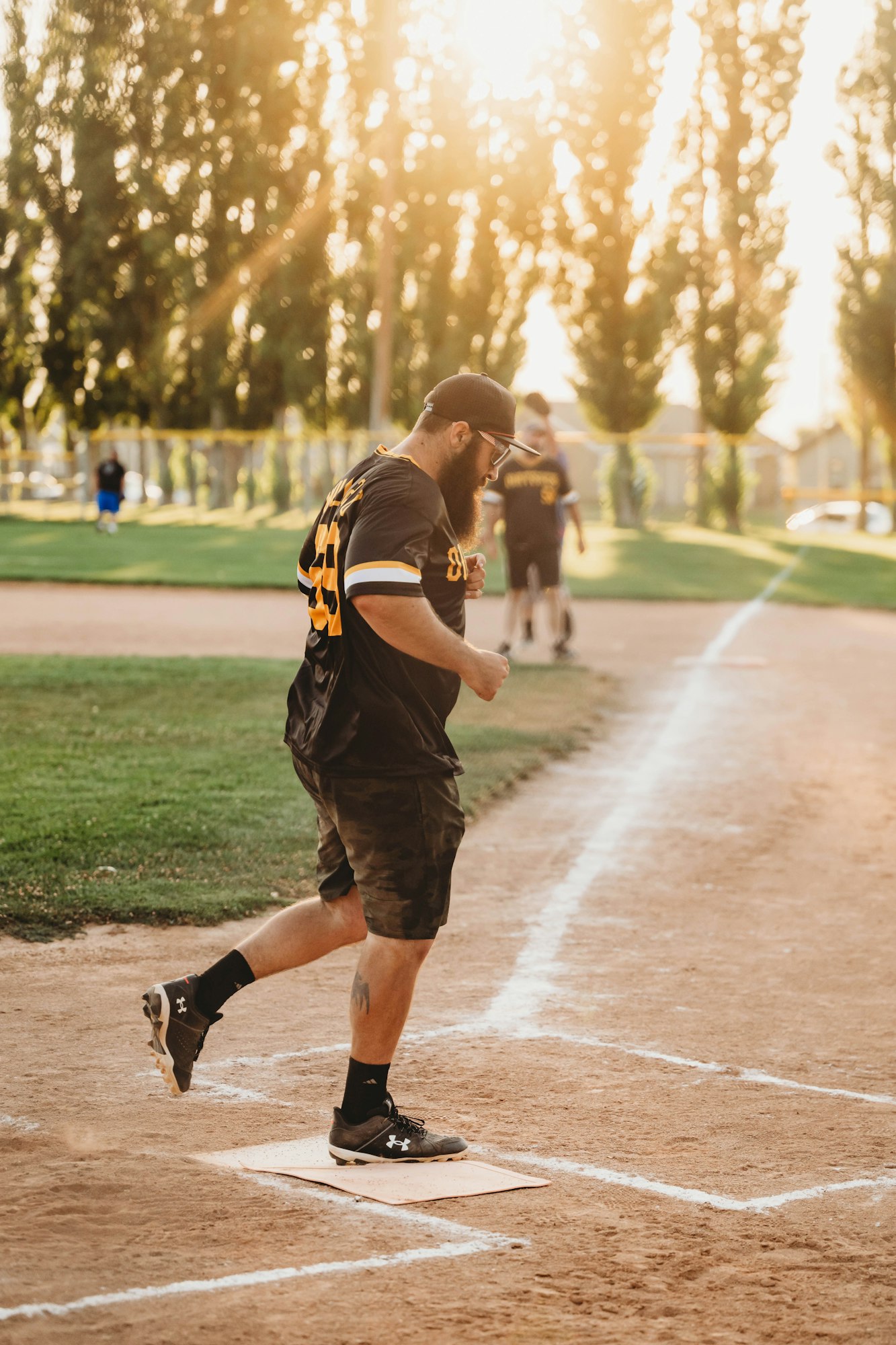 A baseball player stepping on base during a game with sunlight filtering through trees in the background.