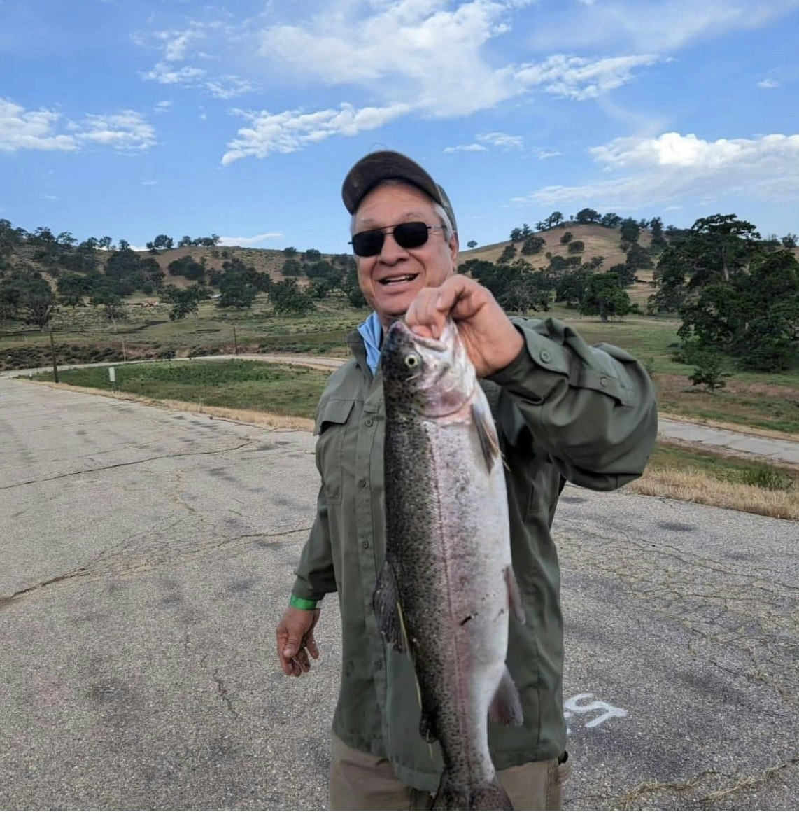 A smiling person holding a fish with a scenic countryside backdrop.