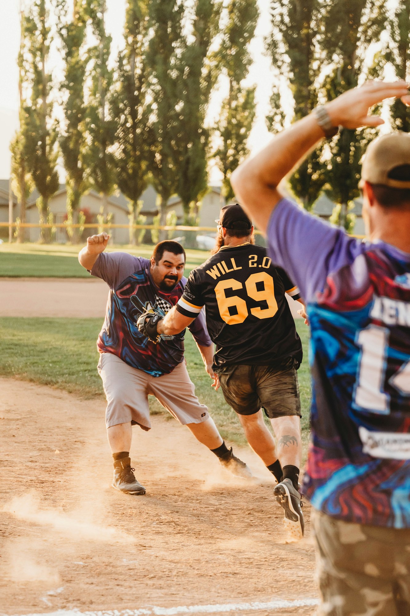 Men playing softball, one in motion to score, dust kicked up, energetic game moment, trees in background.