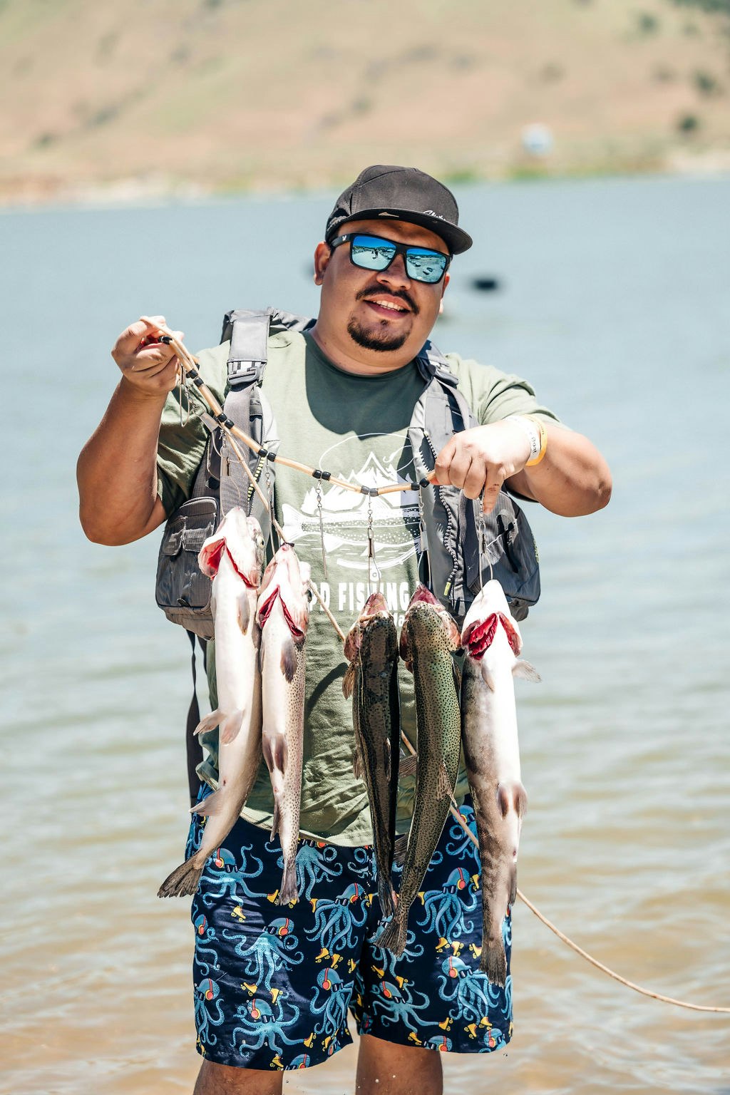 A smiling man in sunglasses stands by water, holding a string of several fish.