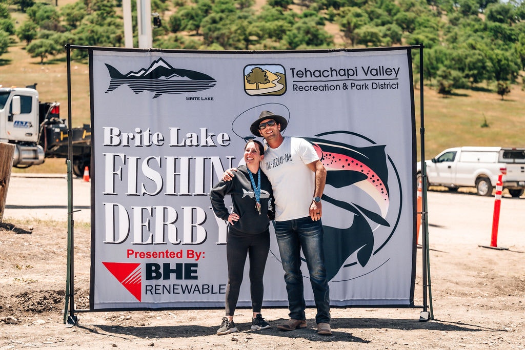 Two people posing by a sign for "Brite Lake Fishing Derby" sponsored by BHE Renewable.