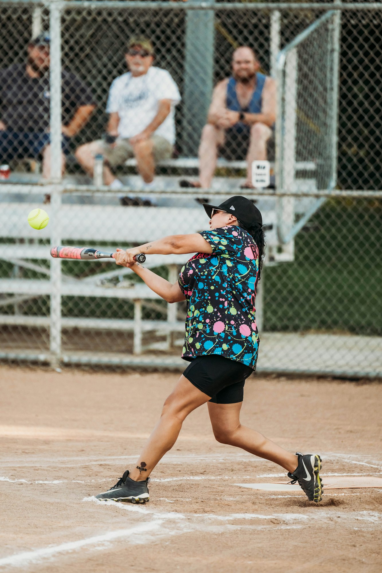 A person swinging a bat at a softball with spectators in the background.