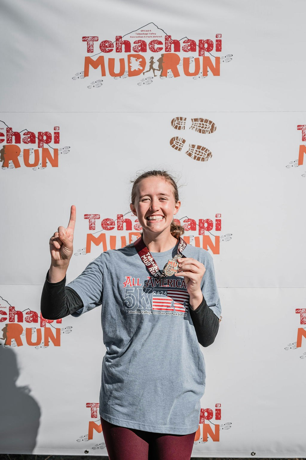 Smiling person holding a medal in front of a "Tehachapi Mud Run" banner.