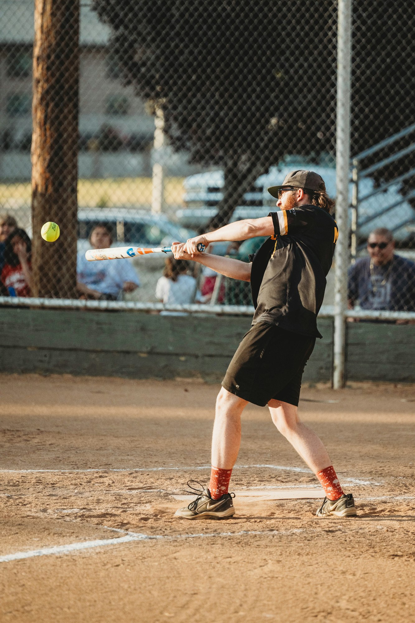 A person playing softball, mid-swing with bat contacting ball, spectators in the background behind a fence.