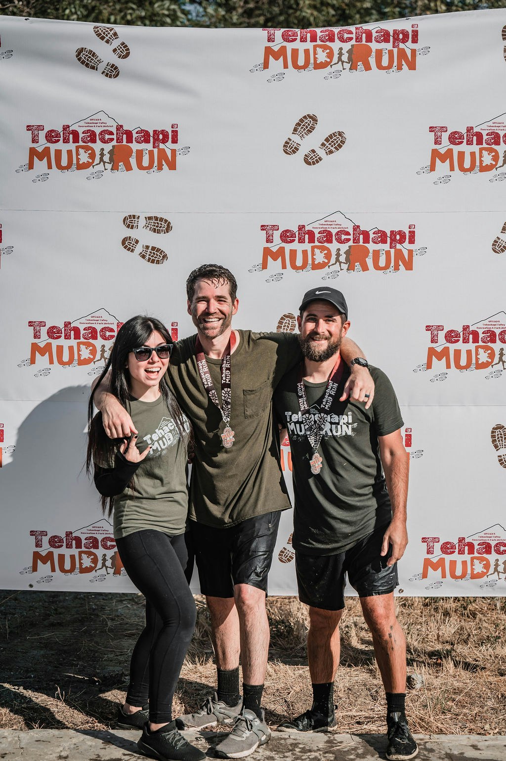 Three people in muddy clothes smiling at the "Tehachapi Mud Run" event backdrop with medals around their necks.