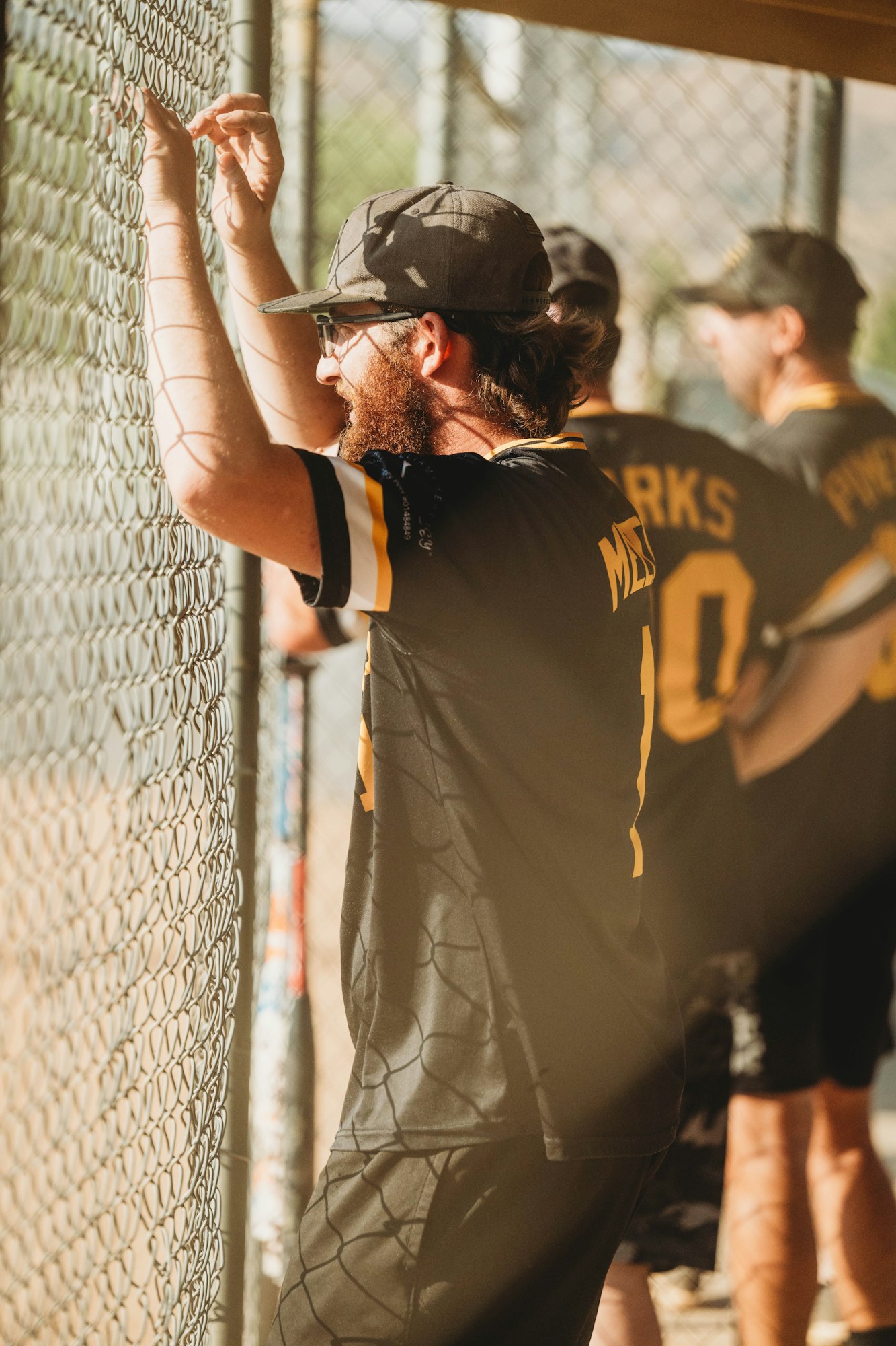 Person in baseball gear leaning on fence, teammates in background, sunny day.