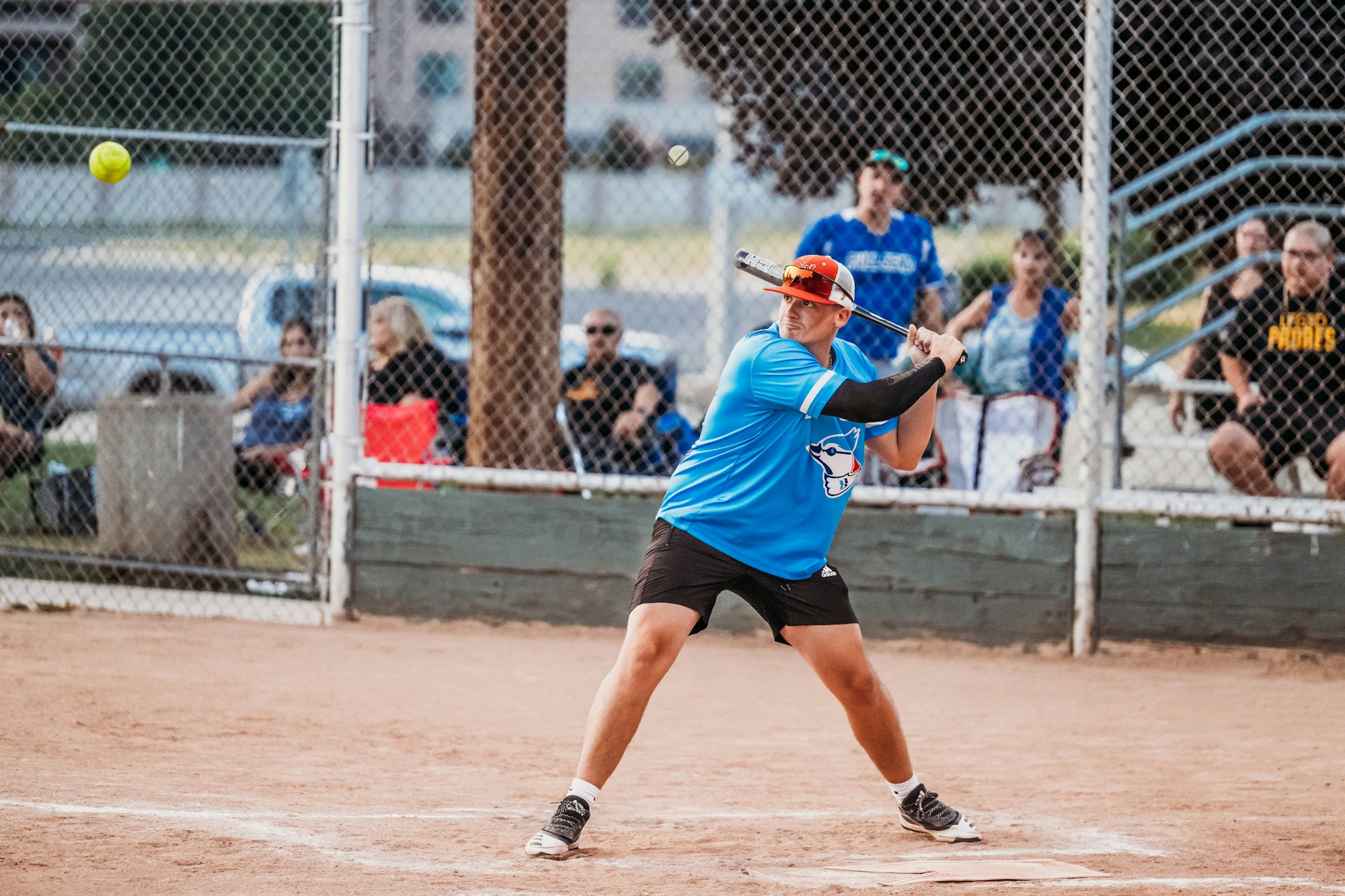 A batter in blue swings at a softball during a game, spectators in the background.