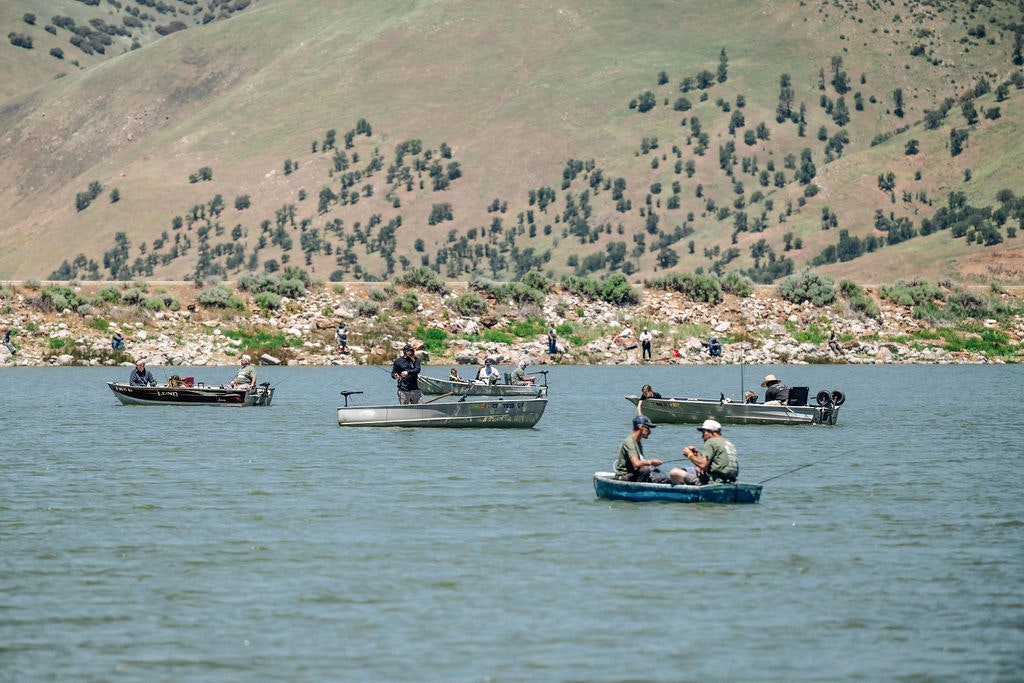 People in boats on a lake with a hilly backdrop. Some appear to be fishing.