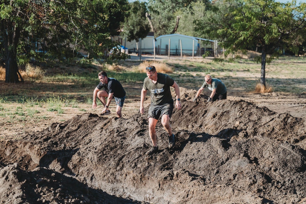 Three people climbing a dirt mound in an outdoor setting with trees and a structure in the background.