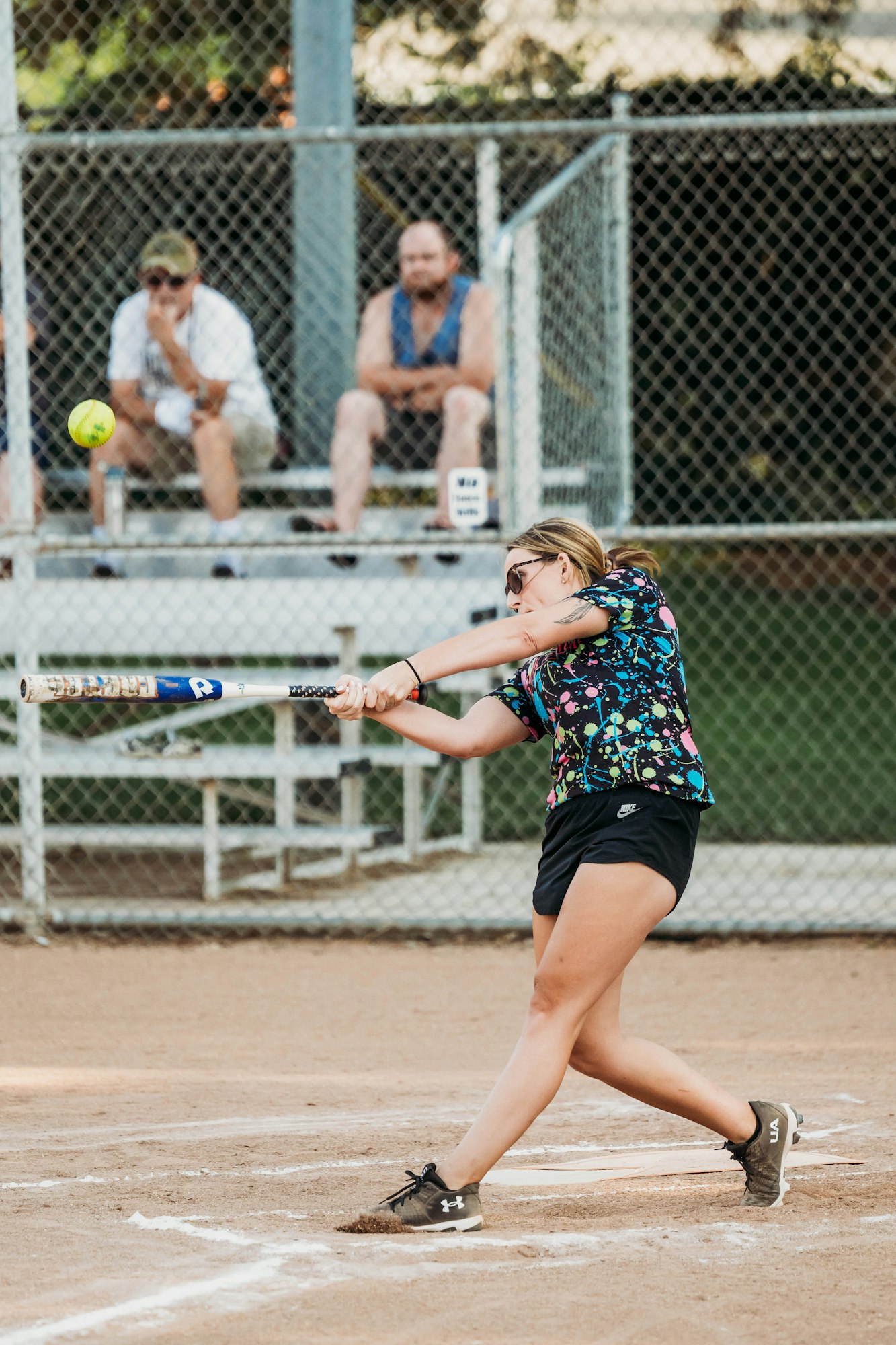 A player swinging at a softball, spectators in the background, on a dirt field.