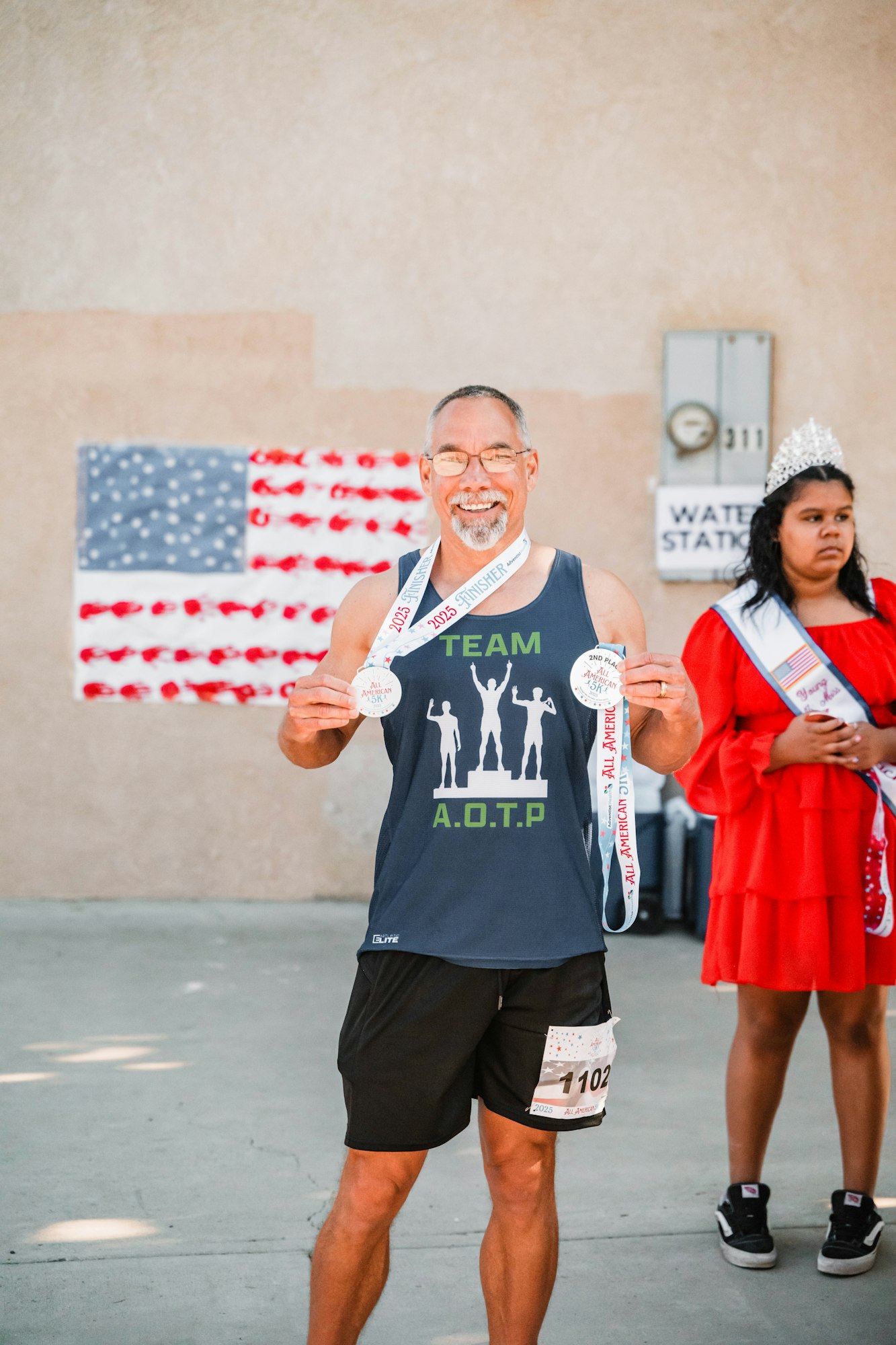 Man smiling with medals, wearing "TEAM A.O.T.P" shirt, next to a woman with a crown. American flag and "WATER STATION" sign in background.