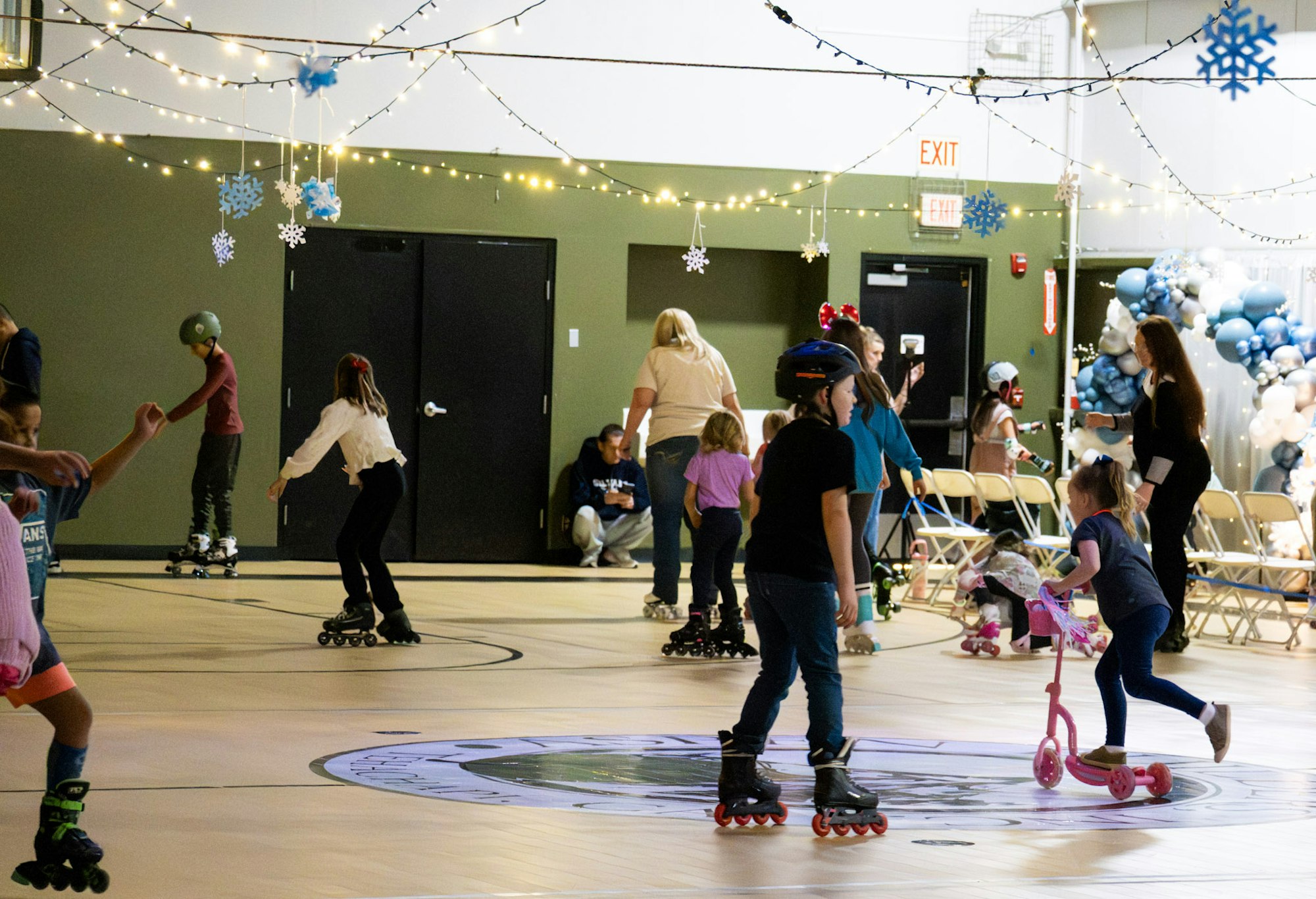 A lively indoor skating rink with kids skating, some on scooters, and festive decorations like lights and snowflakes overhead.