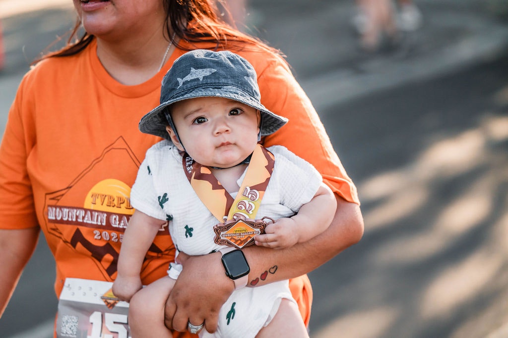 A person in an orange shirt holds a baby wearing a hat and a medal.
